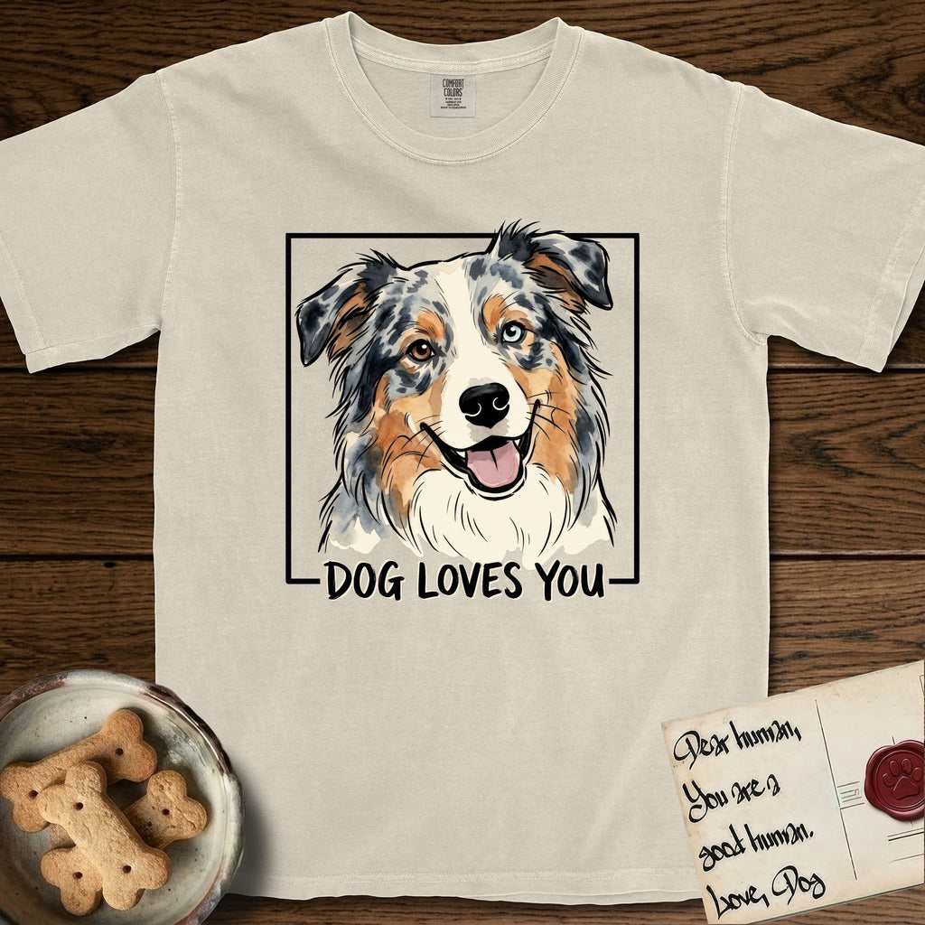 Dog Loves You Australian Shepherd T-Shirt flat lay in Ivory, featuring Ink and watercolor portrait of a smiling blue merle Australian Shepherd with one brown eye and one blue eye and a cream blaze, head tilted inside a black frame above hand-lettered text reading DOG LOVES YOU.