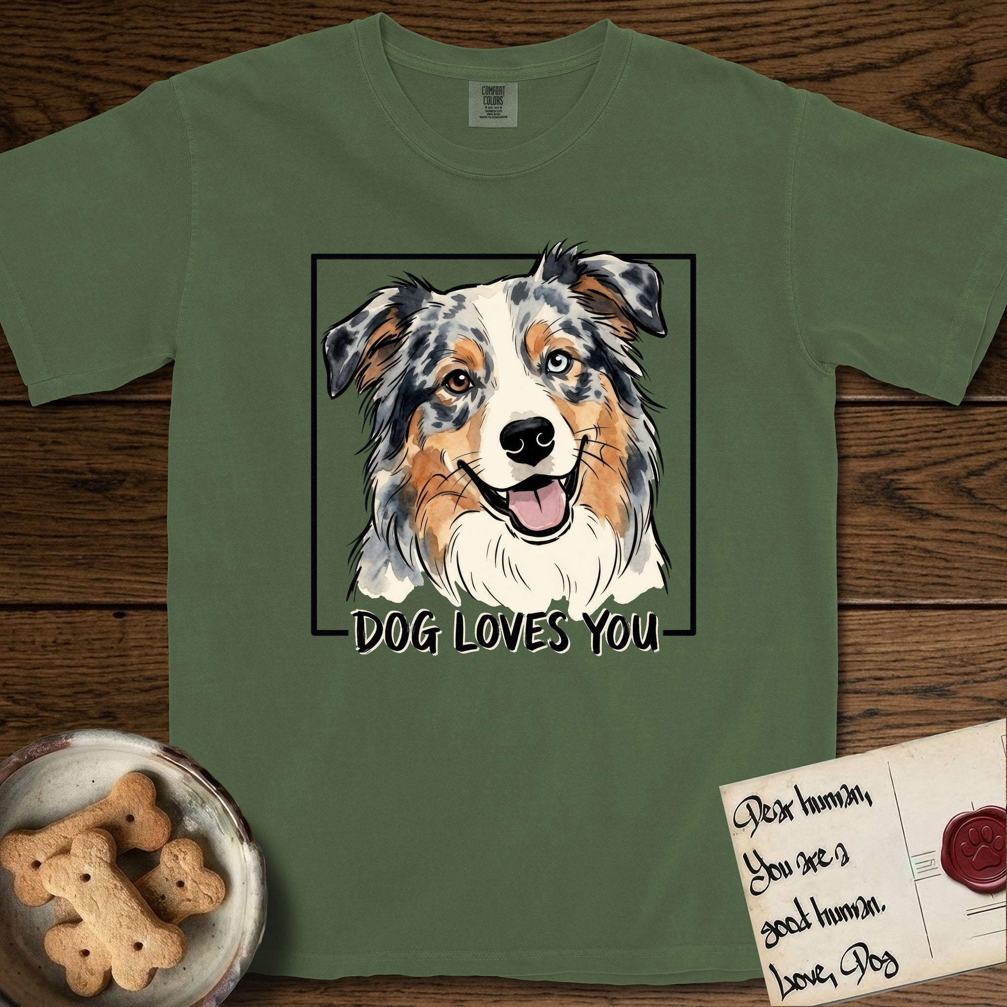 Dog Loves You Australian Shepherd T-Shirt flat lay in Moss, featuring Ink and watercolor portrait of a smiling blue merle Australian Shepherd with one brown eye and one blue eye and a cream blaze, head tilted inside a black frame above hand-lettered text reading DOG LOVES YOU.