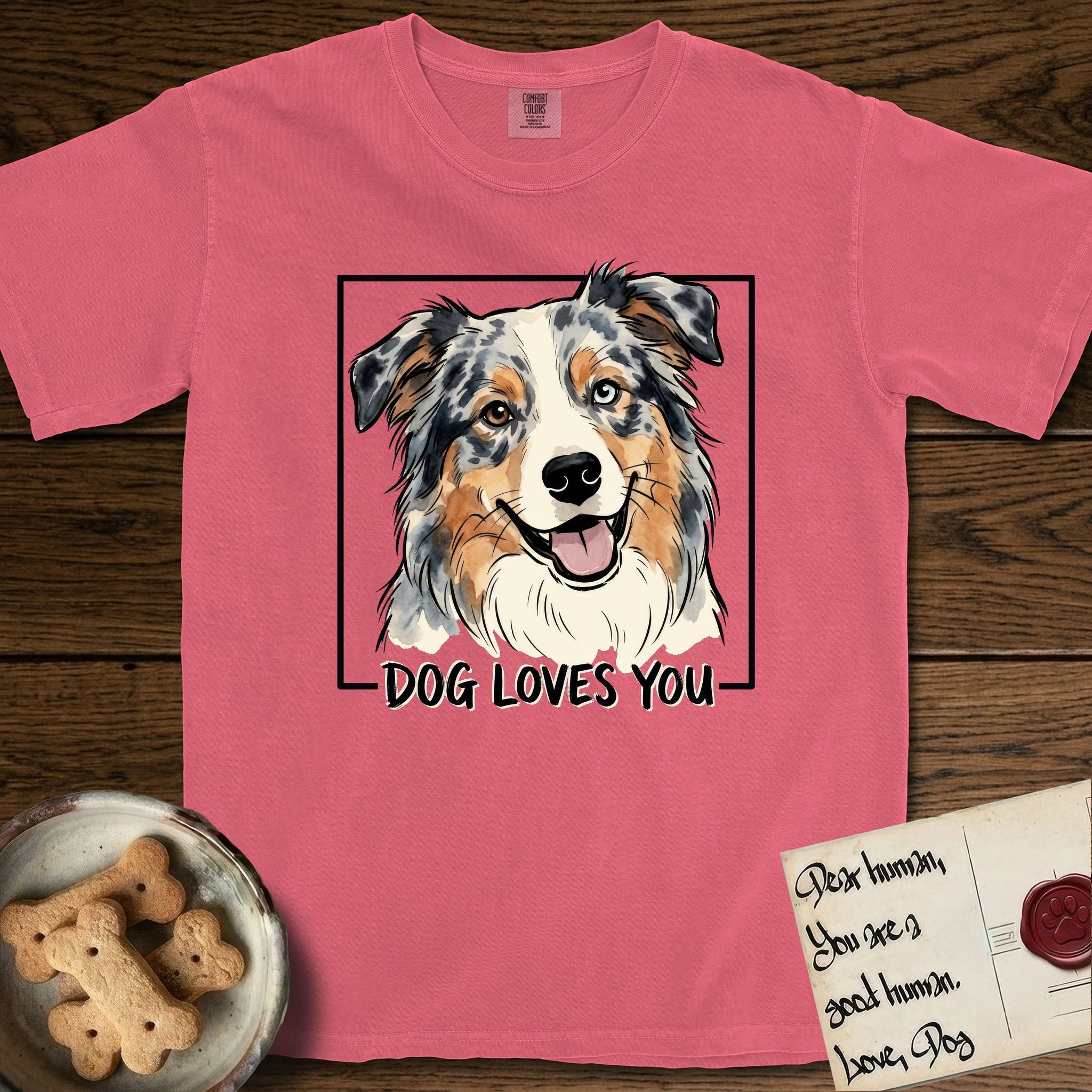 Dog Loves You Australian Shepherd T-Shirt flat lay in Watermelon, featuring Ink and watercolor portrait of a smiling blue merle Australian Shepherd with one brown eye and one blue eye and a cream blaze, head tilted inside a black frame above hand-lettered text reading DOG LOVES YOU.