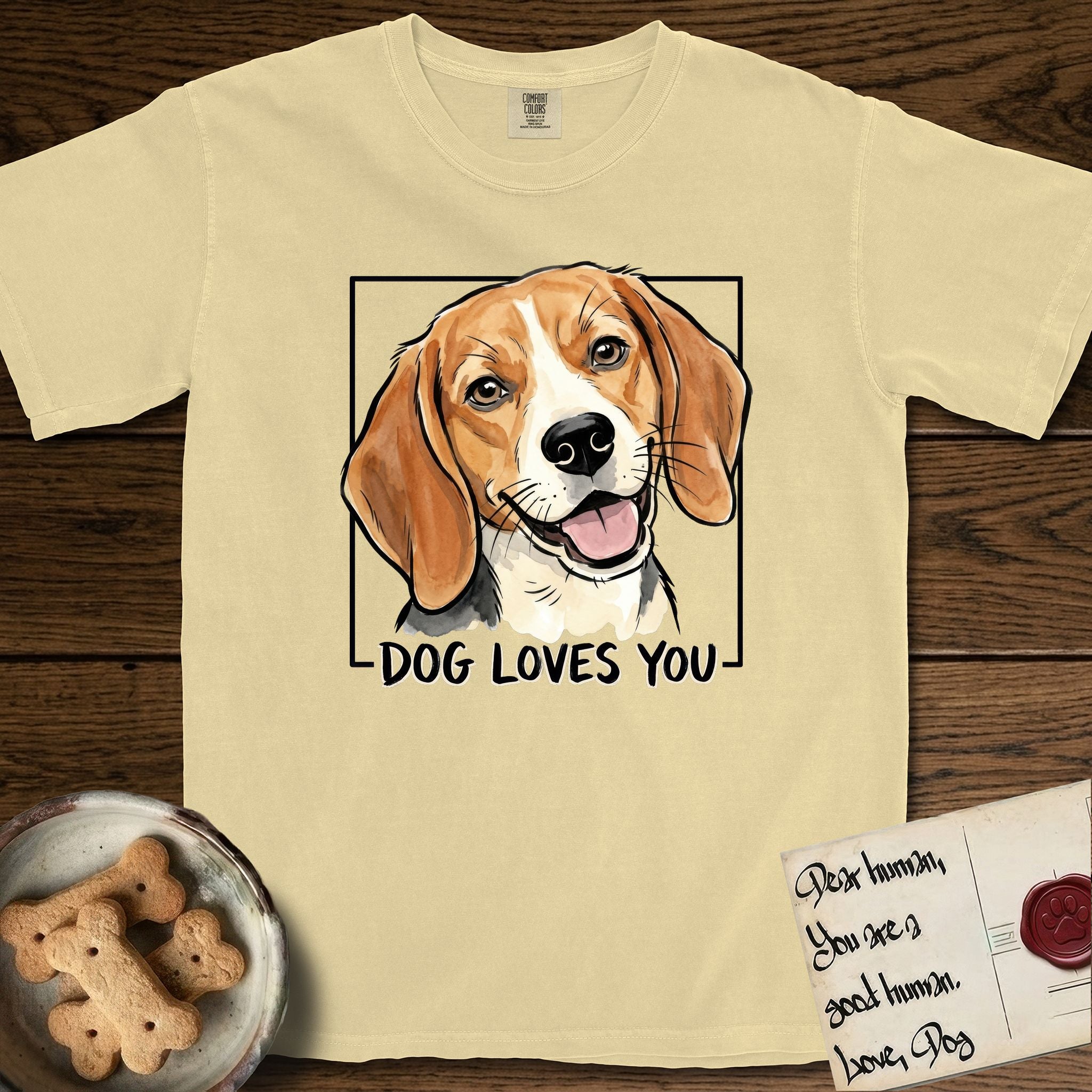 Dog Loves You Beagle T-Shirt flat lay in Butter, featuring Ink and watercolor portrait of a smiling tricolor Beagle with long hanging ears and a cream blaze, head tilted inside a black frame above hand-lettered text reading DOG LOVES YOU.