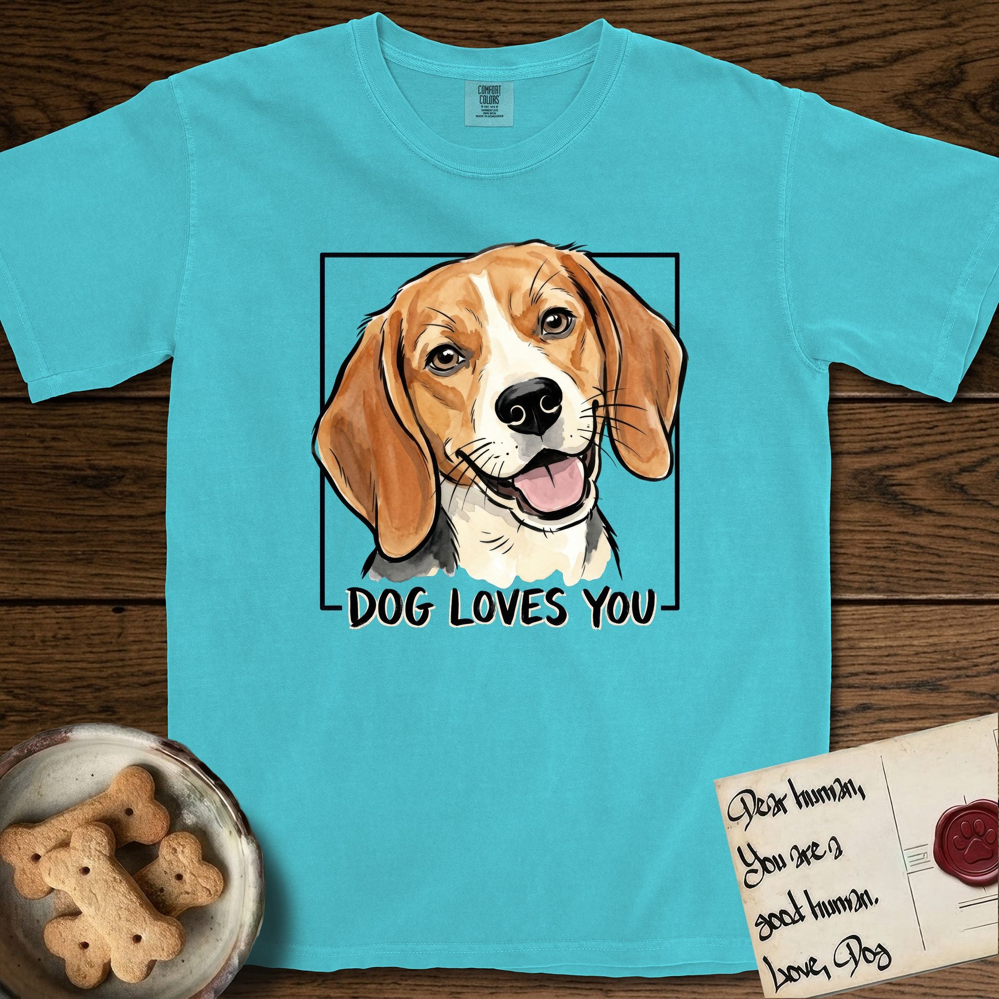 Dog Loves You Beagle T-Shirt flat lay in Lagoon Blue, featuring Ink and watercolor portrait of a smiling tricolor Beagle with long hanging ears and a cream blaze, head tilted inside a black frame above hand-lettered text reading DOG LOVES YOU.