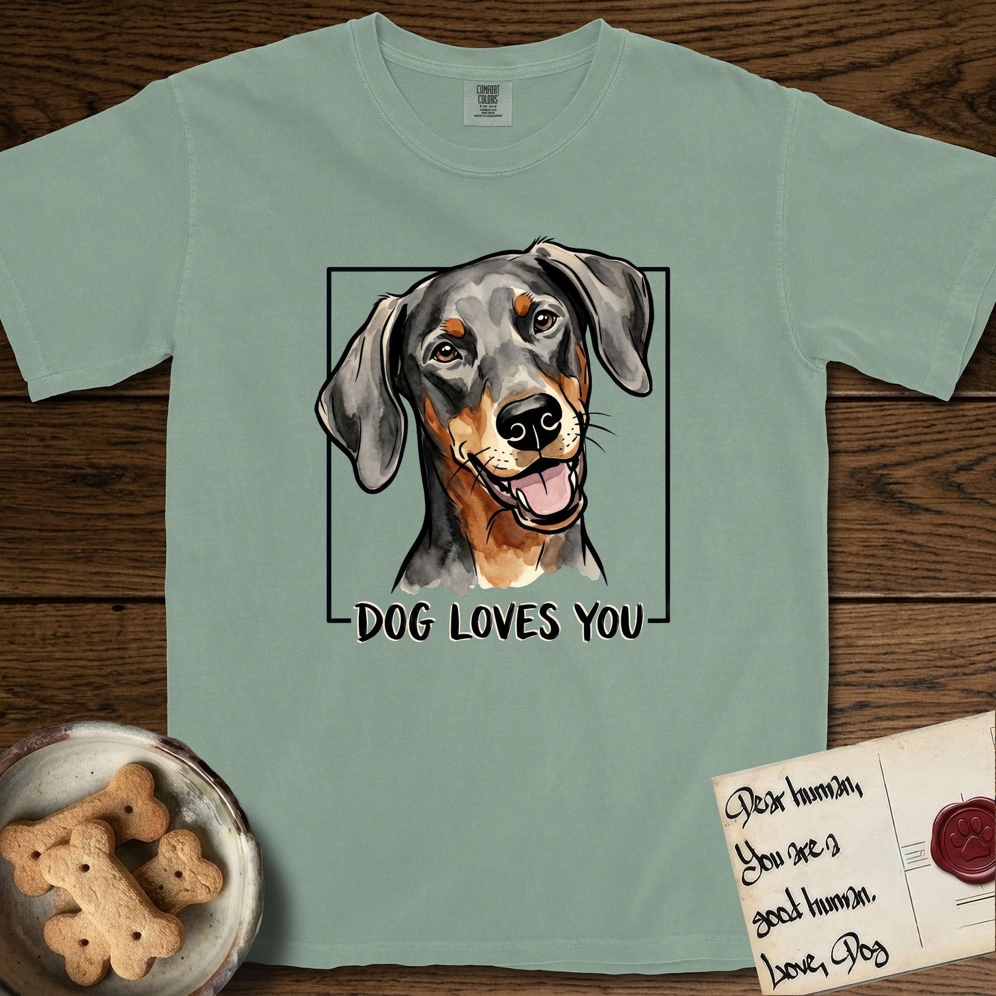 Dog Loves You Doberman T-Shirt flat lay in Bay, featuring Ink and watercolor portrait of a smiling black and rust Doberman with long natural hanging ears, head tilted inside a black frame above hand-lettered text reading DOG LOVES YOU.