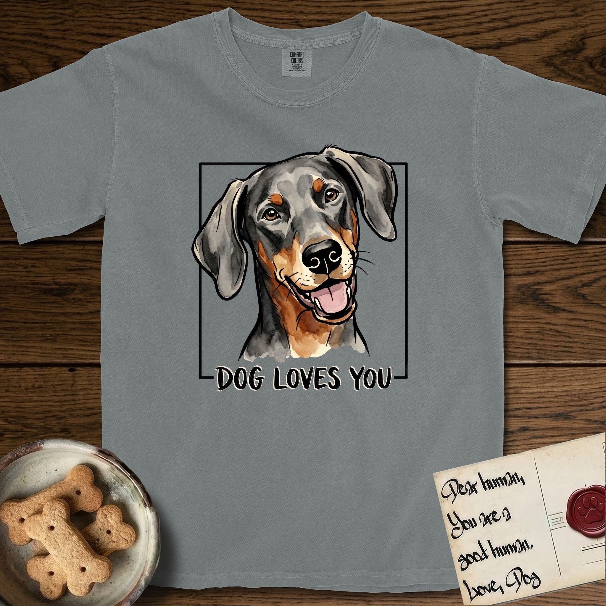 Dog Loves You Doberman T-Shirt flat lay in Granite, featuring Ink and watercolor portrait of a smiling black and rust Doberman with long natural hanging ears, head tilted inside a black frame above hand-lettered text reading DOG LOVES YOU.