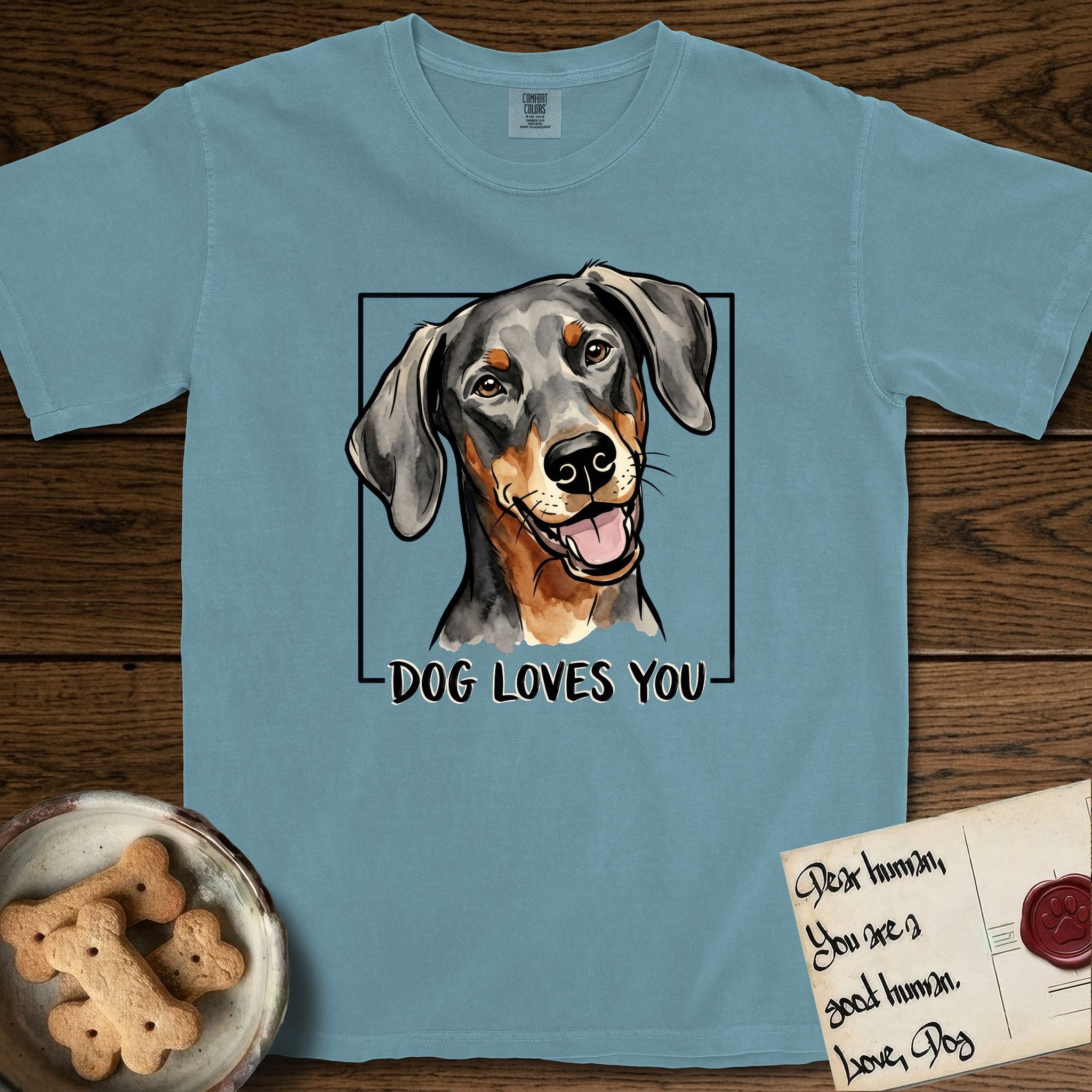 Dog Loves You Doberman T-Shirt flat lay in Ice Blue, featuring Ink and watercolor portrait of a smiling black and rust Doberman with long natural hanging ears, head tilted inside a black frame above hand-lettered text reading DOG LOVES YOU.