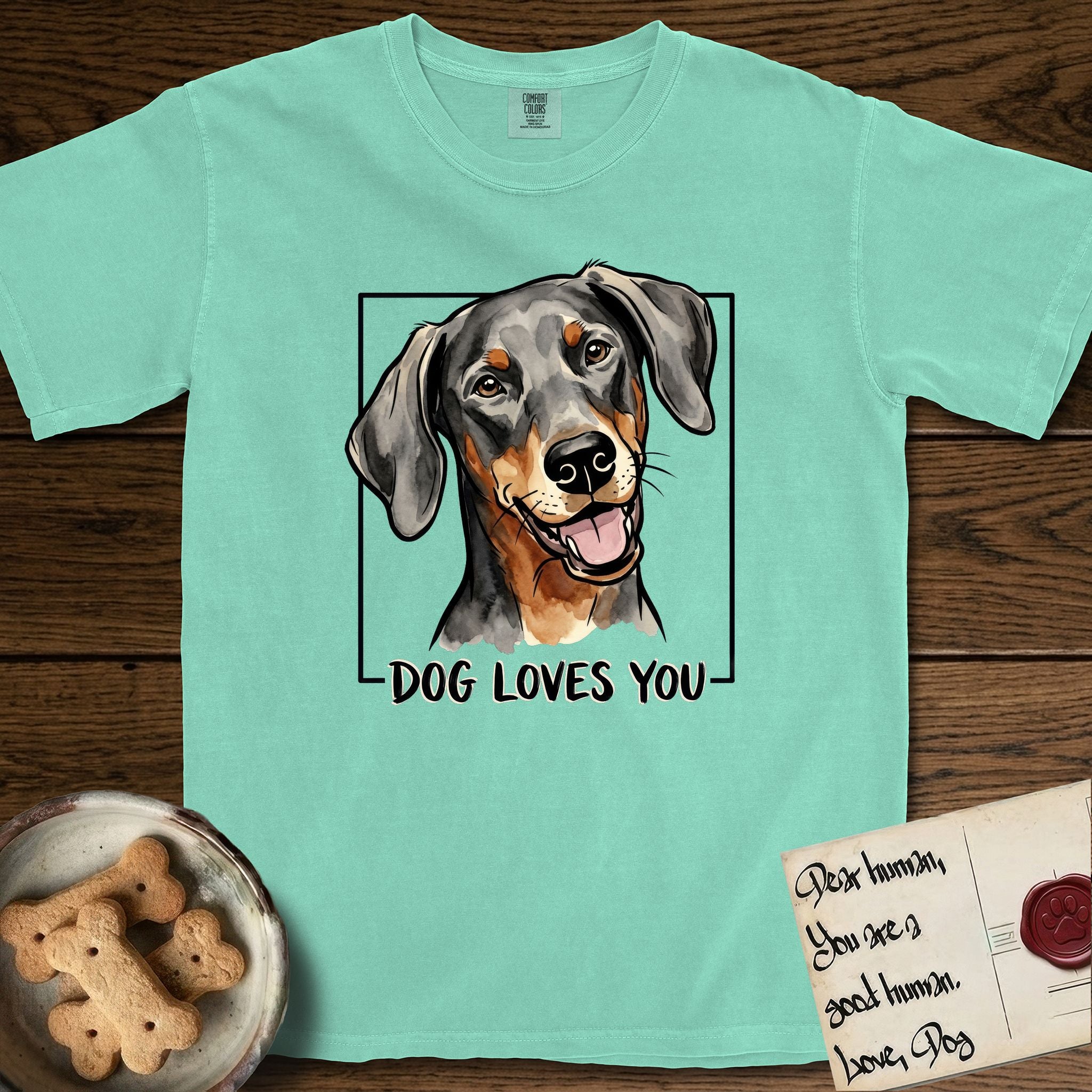 Dog Loves You Doberman T-Shirt flat lay in Island Reef, featuring Ink and watercolor portrait of a smiling black and rust Doberman with long natural hanging ears, head tilted inside a black frame above hand-lettered text reading DOG LOVES YOU.