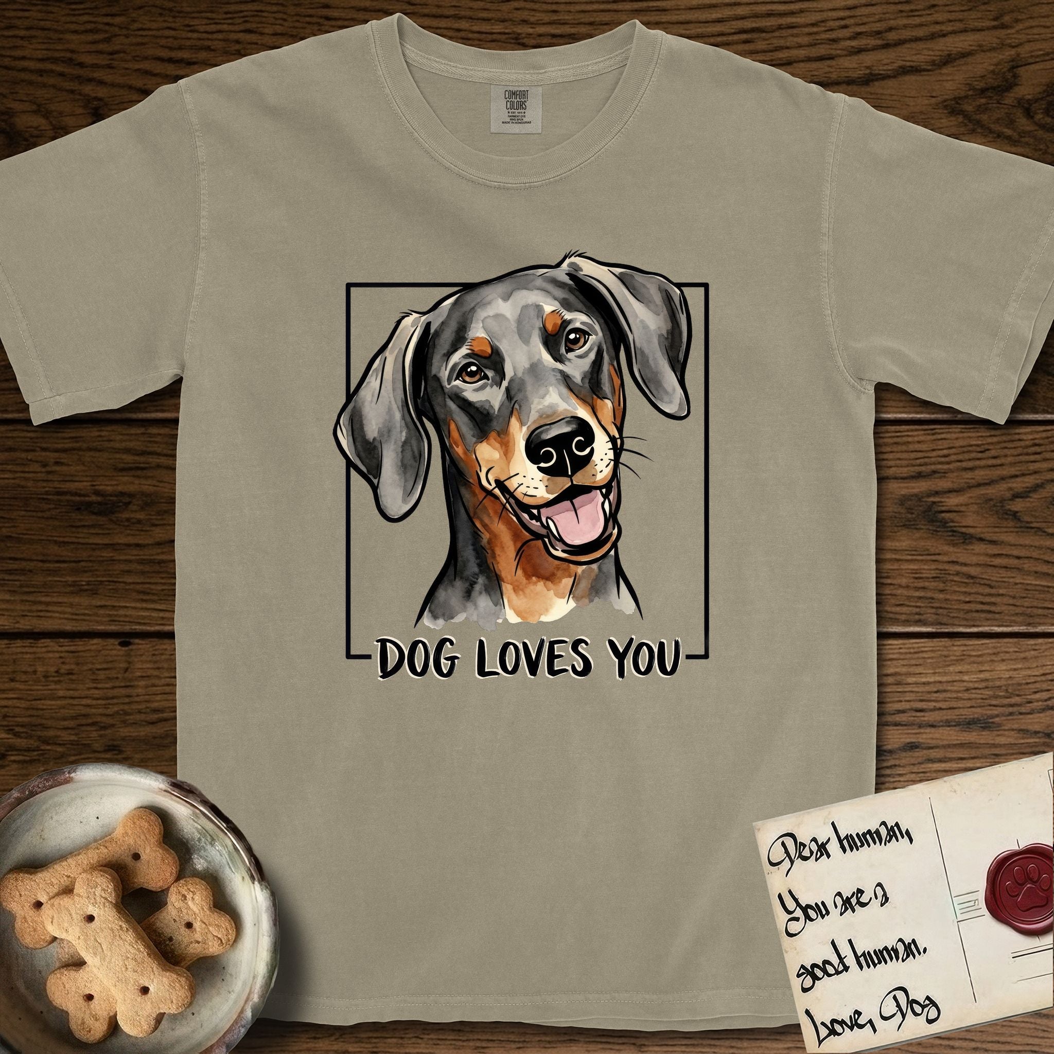 Dog Loves You Doberman T-Shirt flat lay in Khaki, featuring Ink and watercolor portrait of a smiling black and rust Doberman with long natural hanging ears, head tilted inside a black frame above hand-lettered text reading DOG LOVES YOU.