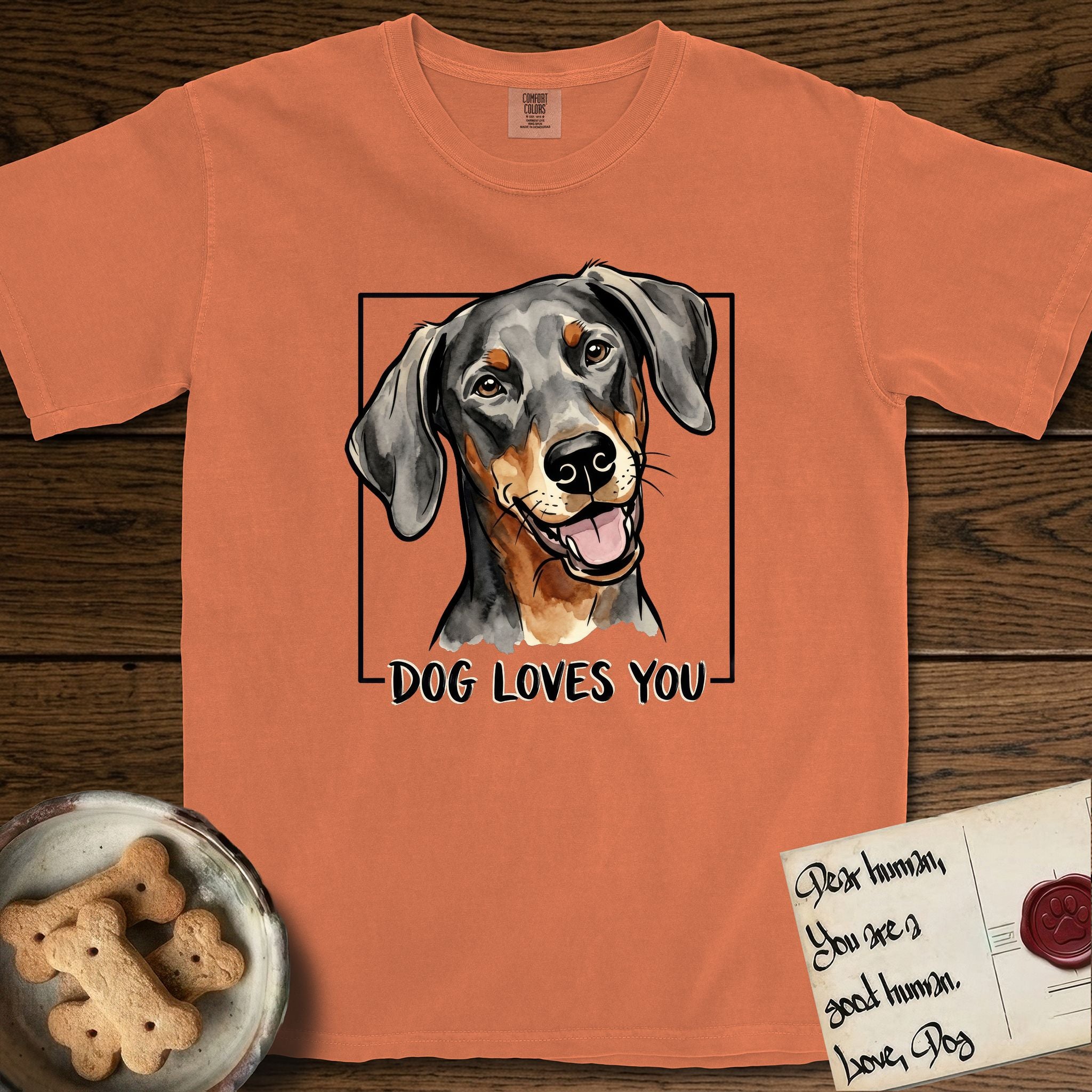 Dog Loves You Doberman T-Shirt flat lay in Melon, featuring Ink and watercolor portrait of a smiling black and rust Doberman with long natural hanging ears, head tilted inside a black frame above hand-lettered text reading DOG LOVES YOU.