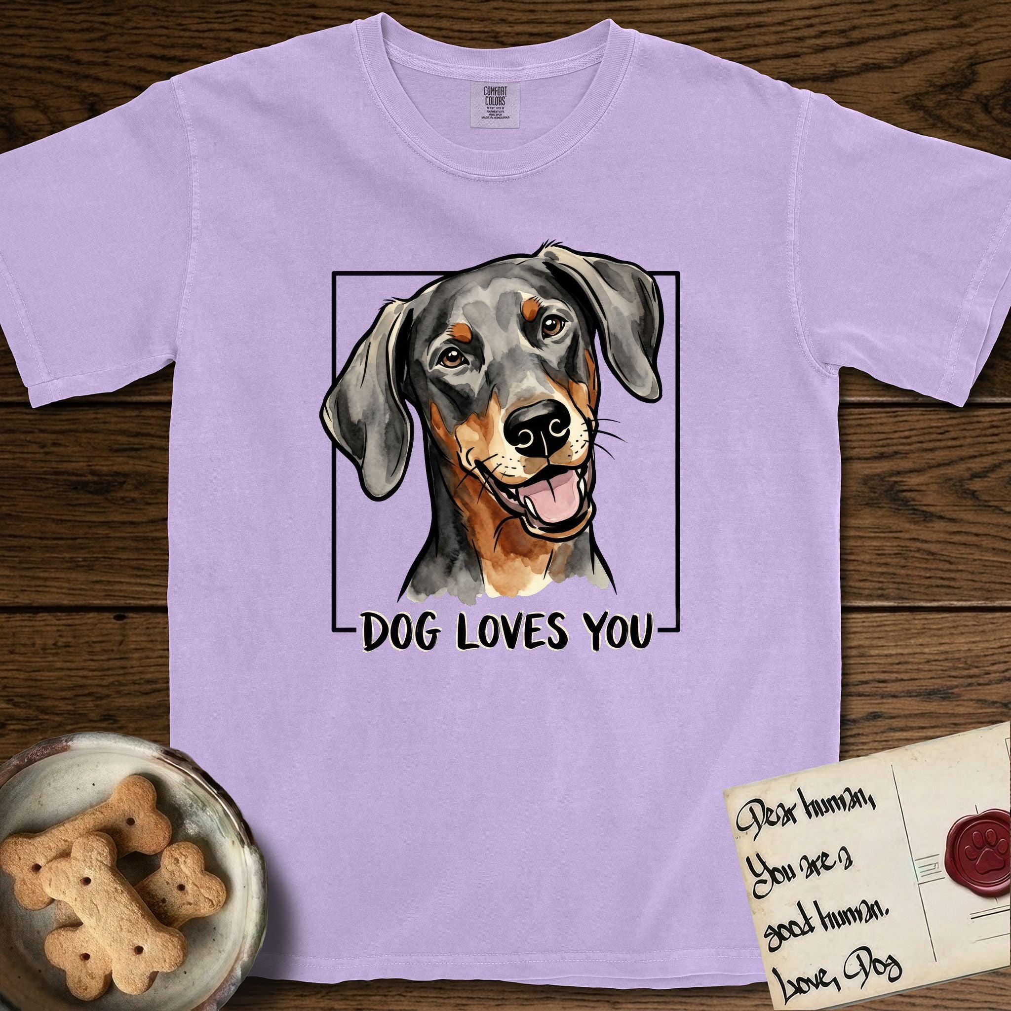 Dog Loves You Doberman T-Shirt flat lay in Orchid, featuring Ink and watercolor portrait of a smiling black and rust Doberman with long natural hanging ears, head tilted inside a black frame above hand-lettered text reading DOG LOVES YOU.