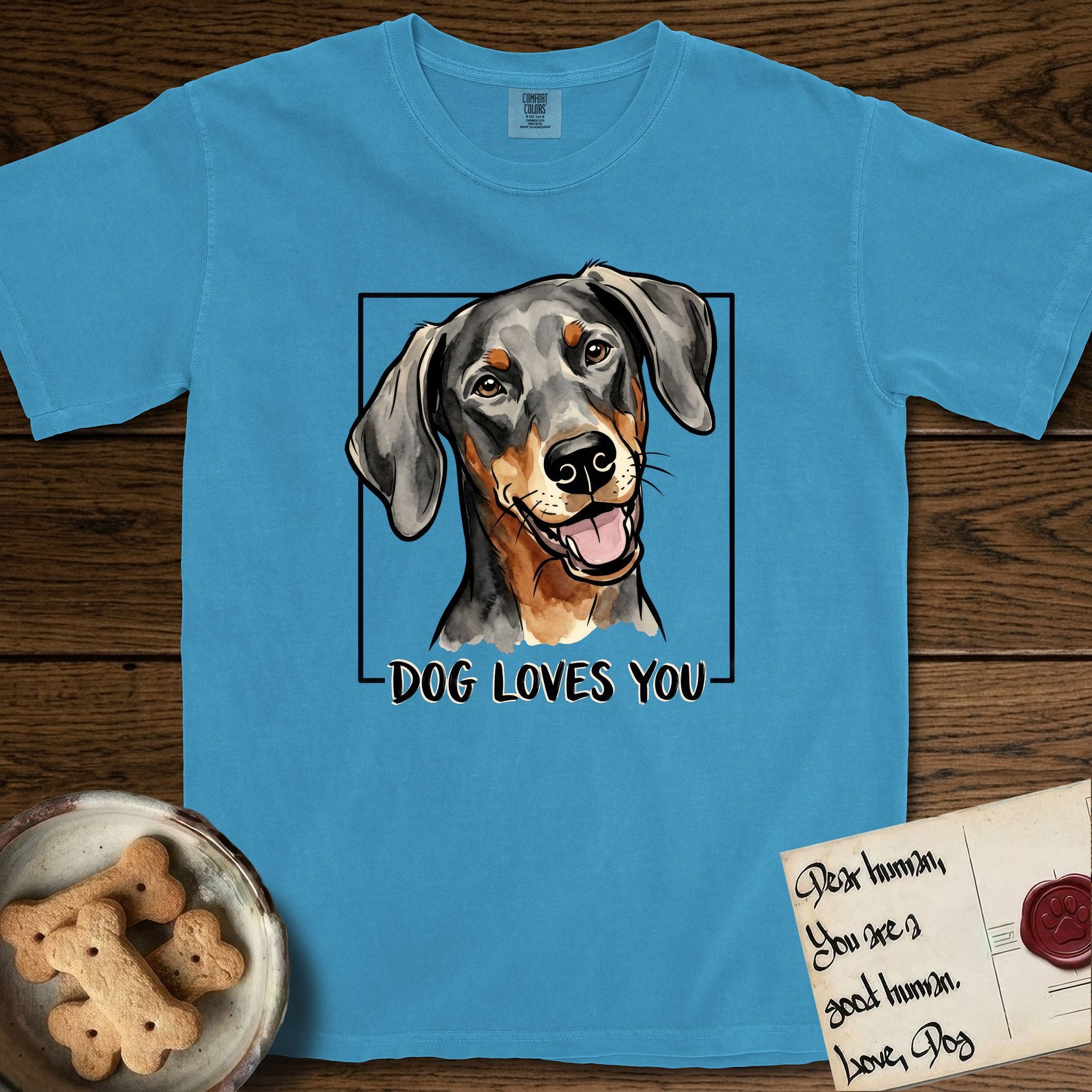 Dog Loves You Doberman T-Shirt flat lay in Royal Caribe, featuring Ink and watercolor portrait of a smiling black and rust Doberman with long natural hanging ears, head tilted inside a black frame above hand-lettered text reading DOG LOVES YOU.