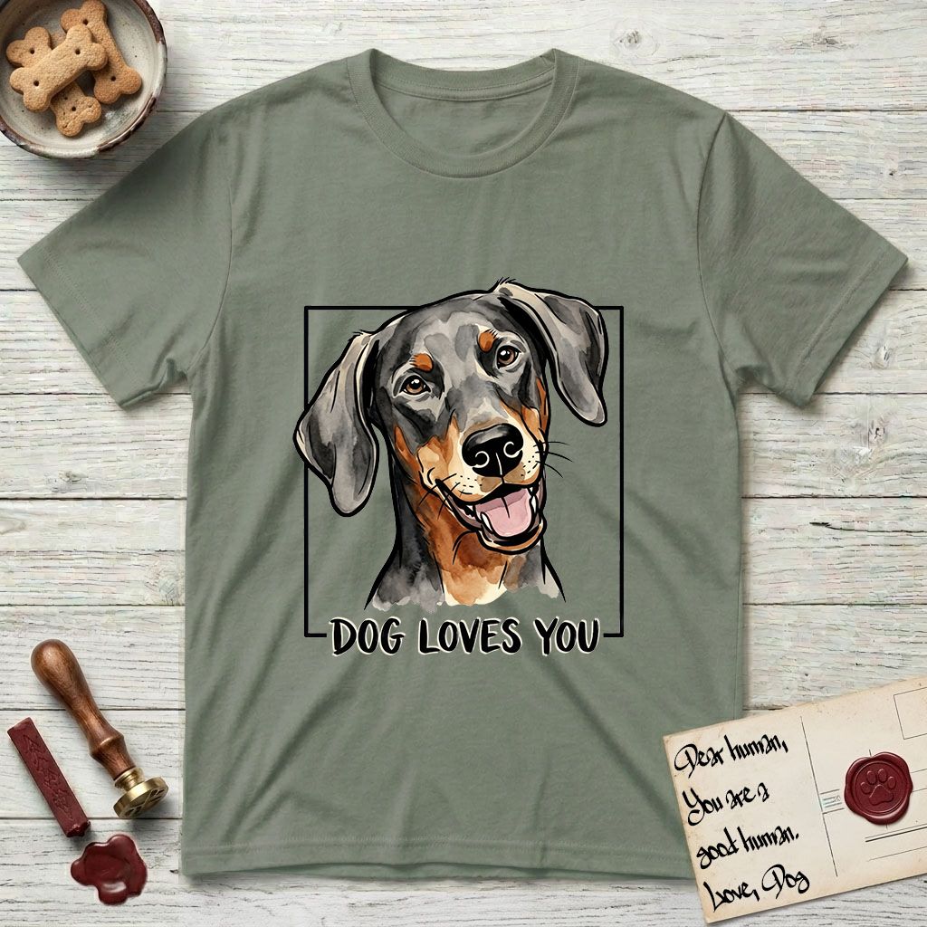 Dog Loves You Doberman T-Shirt flat lay in Sage, featuring Ink and watercolor portrait of a smiling black and rust Doberman with long natural hanging ears, head tilted inside a black frame above hand-lettered text reading DOG LOVES YOU.