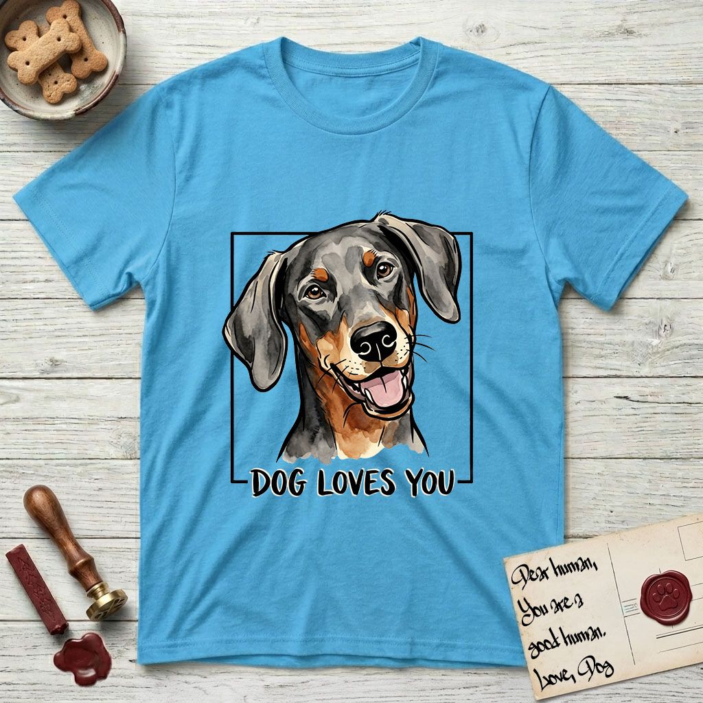 Dog Loves You Doberman T-Shirt flat lay in Sky, featuring Ink and watercolor portrait of a smiling black and rust Doberman with long natural hanging ears, head tilted inside a black frame above hand-lettered text reading DOG LOVES YOU.