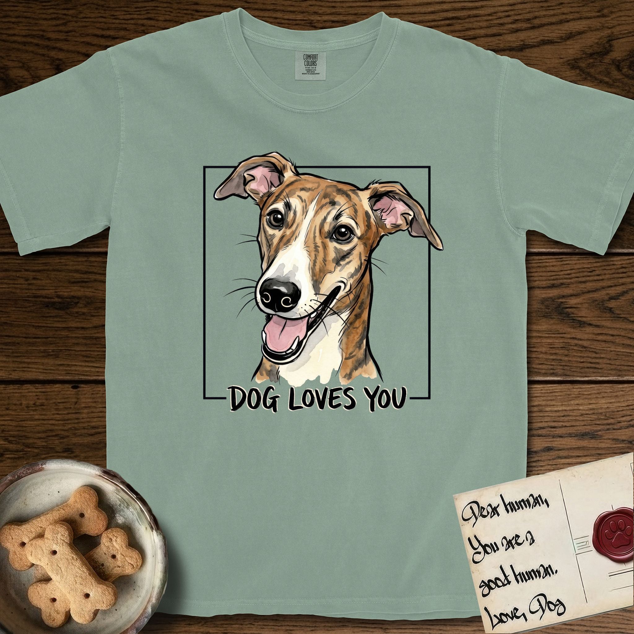 Dog Loves You Galgo T-Shirt flat lay in Bay, featuring Ink and watercolor portrait of a smiling brindle Galgo with a long narrow muzzle and folded rose ears, head tilted inside a black frame above hand-lettered text reading DOG LOVES YOU.