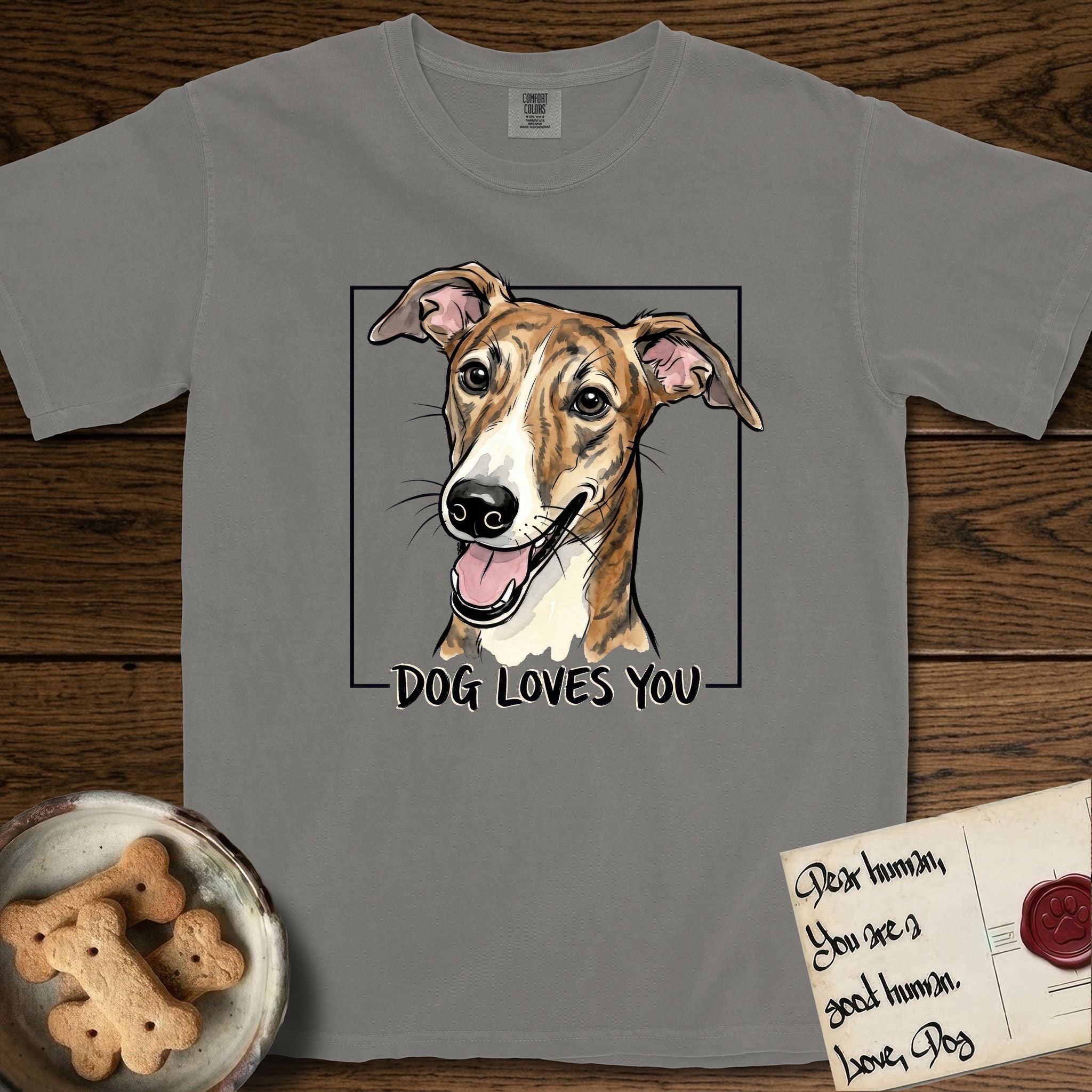 Dog Loves You Galgo T-Shirt flat lay in Grey, featuring Ink and watercolor portrait of a smiling brindle Galgo with a long narrow muzzle and folded rose ears, head tilted inside a black frame above hand-lettered text reading DOG LOVES YOU.
