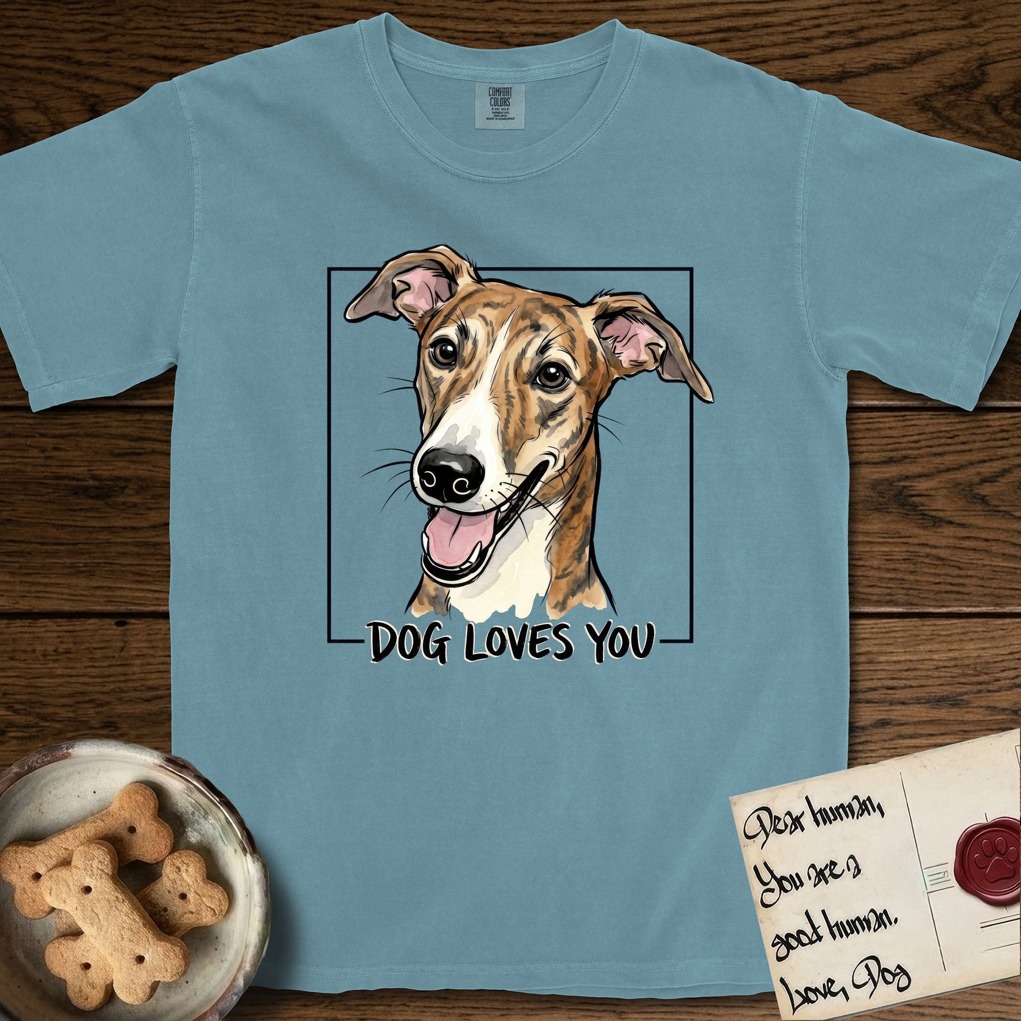 Dog Loves You Galgo T-Shirt flat lay in Ice Blue, featuring Ink and watercolor portrait of a smiling brindle Galgo with a long narrow muzzle and folded rose ears, head tilted inside a black frame above hand-lettered text reading DOG LOVES YOU.