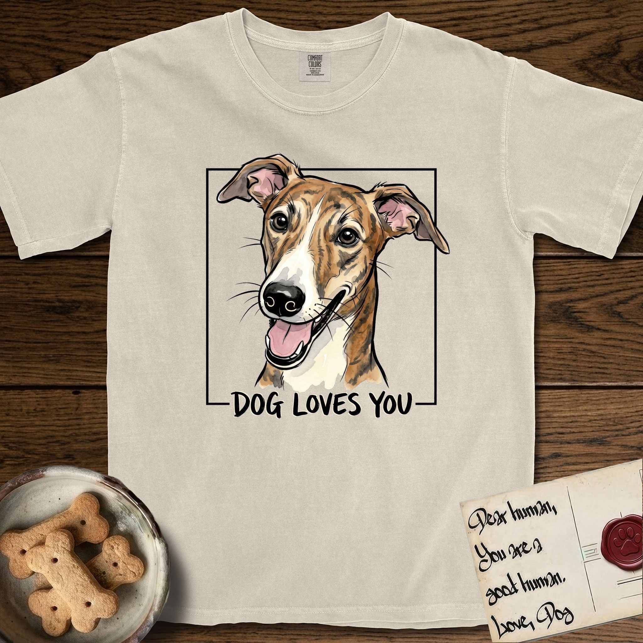 Dog Loves You Galgo T-Shirt flat lay in Ivory, featuring Ink and watercolor portrait of a smiling brindle Galgo with a long narrow muzzle and folded rose ears, head tilted inside a black frame above hand-lettered text reading DOG LOVES YOU.