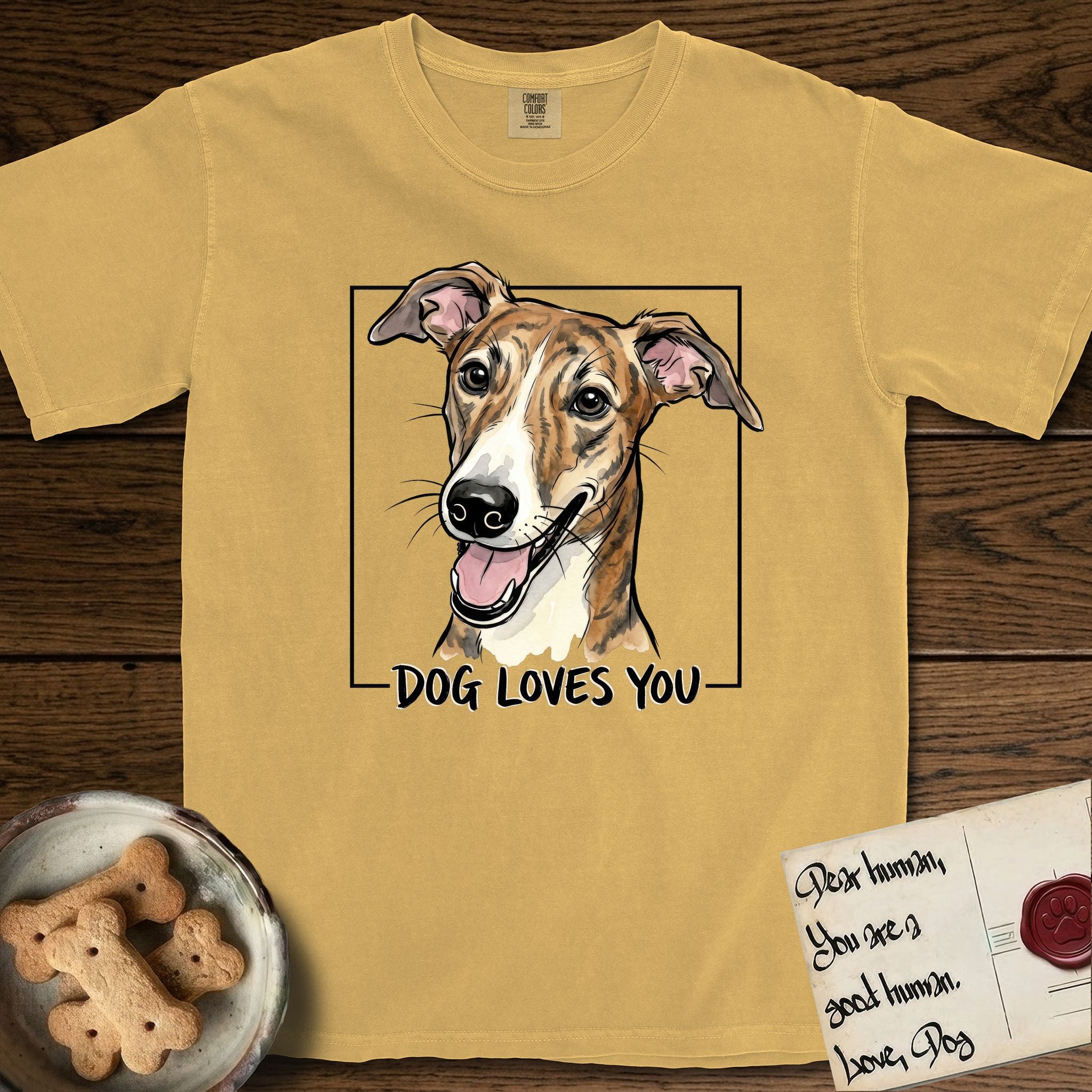 Dog Loves You Galgo T-Shirt flat lay in Mustard, featuring Ink and watercolor portrait of a smiling brindle Galgo with a long narrow muzzle and folded rose ears, head tilted inside a black frame above hand-lettered text reading DOG LOVES YOU.
