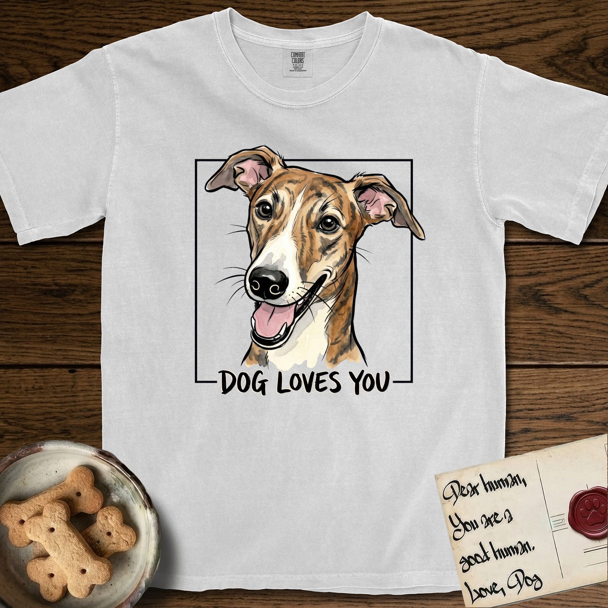 Dog Loves You Galgo T-Shirt flat lay in White, featuring Ink and watercolor portrait of a smiling brindle Galgo with a long narrow muzzle and folded rose ears, head tilted inside a black frame above hand-lettered text reading DOG LOVES YOU.