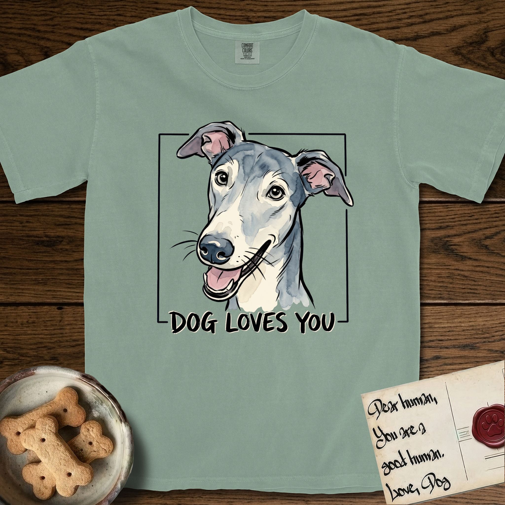Dog Loves You Greyhound T-Shirt flat lay in Bay, featuring Ink and watercolor portrait of a smiling slate blue Greyhound with a long narrow muzzle, cream blaze and folded rose ears, head tilted inside a black frame above hand-lettered text reading DOG LOVES YOU.