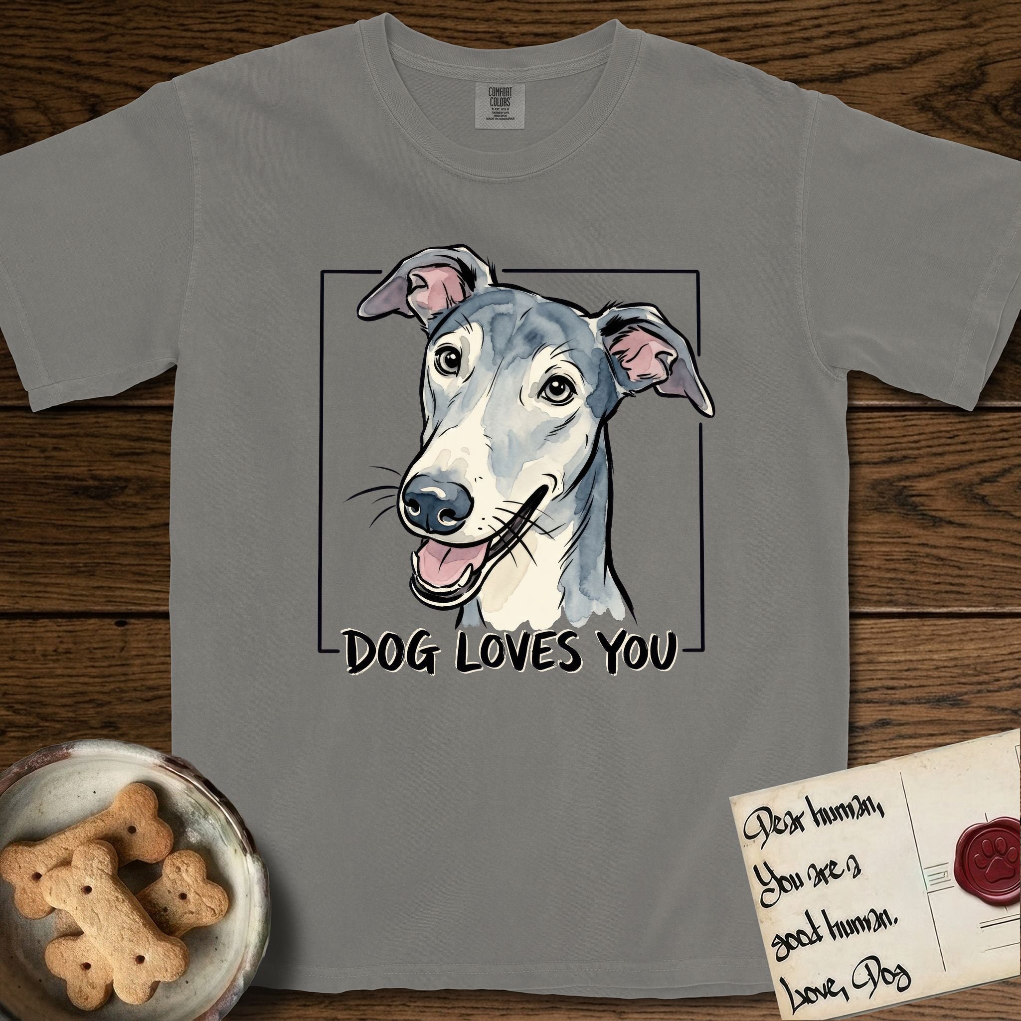 Dog Loves You Greyhound T-Shirt flat lay in Grey, featuring Ink and watercolor portrait of a smiling slate blue Greyhound with a long narrow muzzle, cream blaze and folded rose ears, head tilted inside a black frame above hand-lettered text reading DOG LOVES YOU.