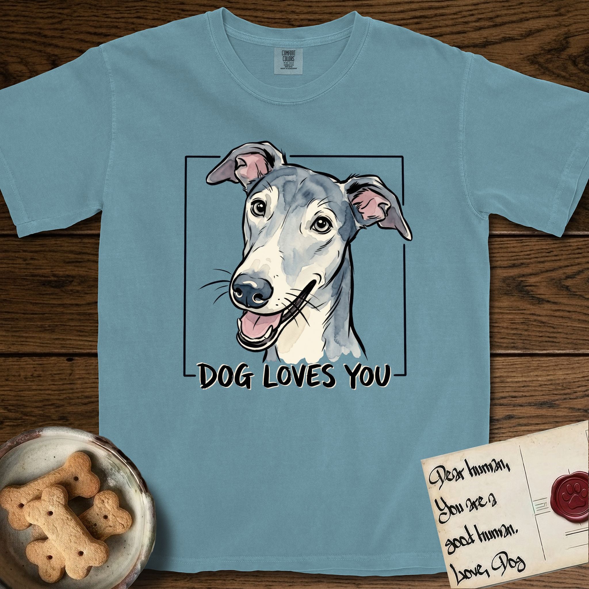 Dog Loves You Greyhound T-Shirt flat lay in Ice Blue, featuring Ink and watercolor portrait of a smiling slate blue Greyhound with a long narrow muzzle, cream blaze and folded rose ears, head tilted inside a black frame above hand-lettered text reading DOG LOVES YOU.