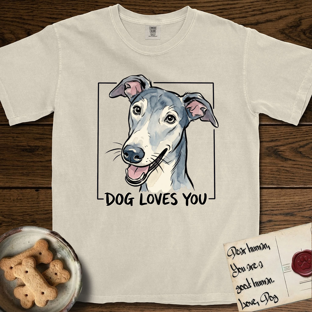 Dog Loves You Greyhound T-Shirt flat lay in Ivory, featuring Ink and watercolor portrait of a smiling slate blue Greyhound with a long narrow muzzle, cream blaze and folded rose ears, head tilted inside a black frame above hand-lettered text reading DOG LOVES YOU.