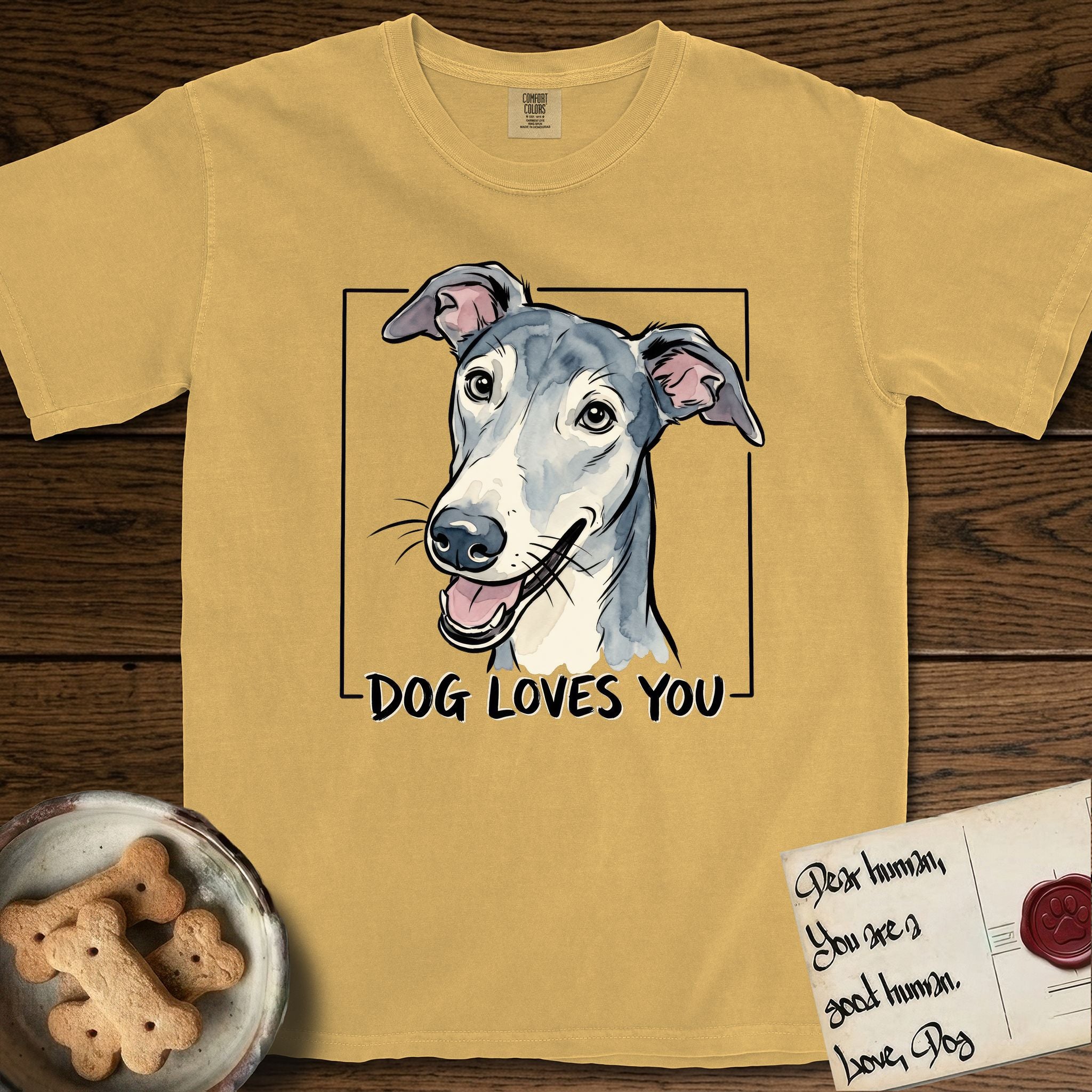 Dog Loves You Greyhound T-Shirt flat lay in Mustard, featuring Ink and watercolor portrait of a smiling slate blue Greyhound with a long narrow muzzle, cream blaze and folded rose ears, head tilted inside a black frame above hand-lettered text reading DOG LOVES YOU.