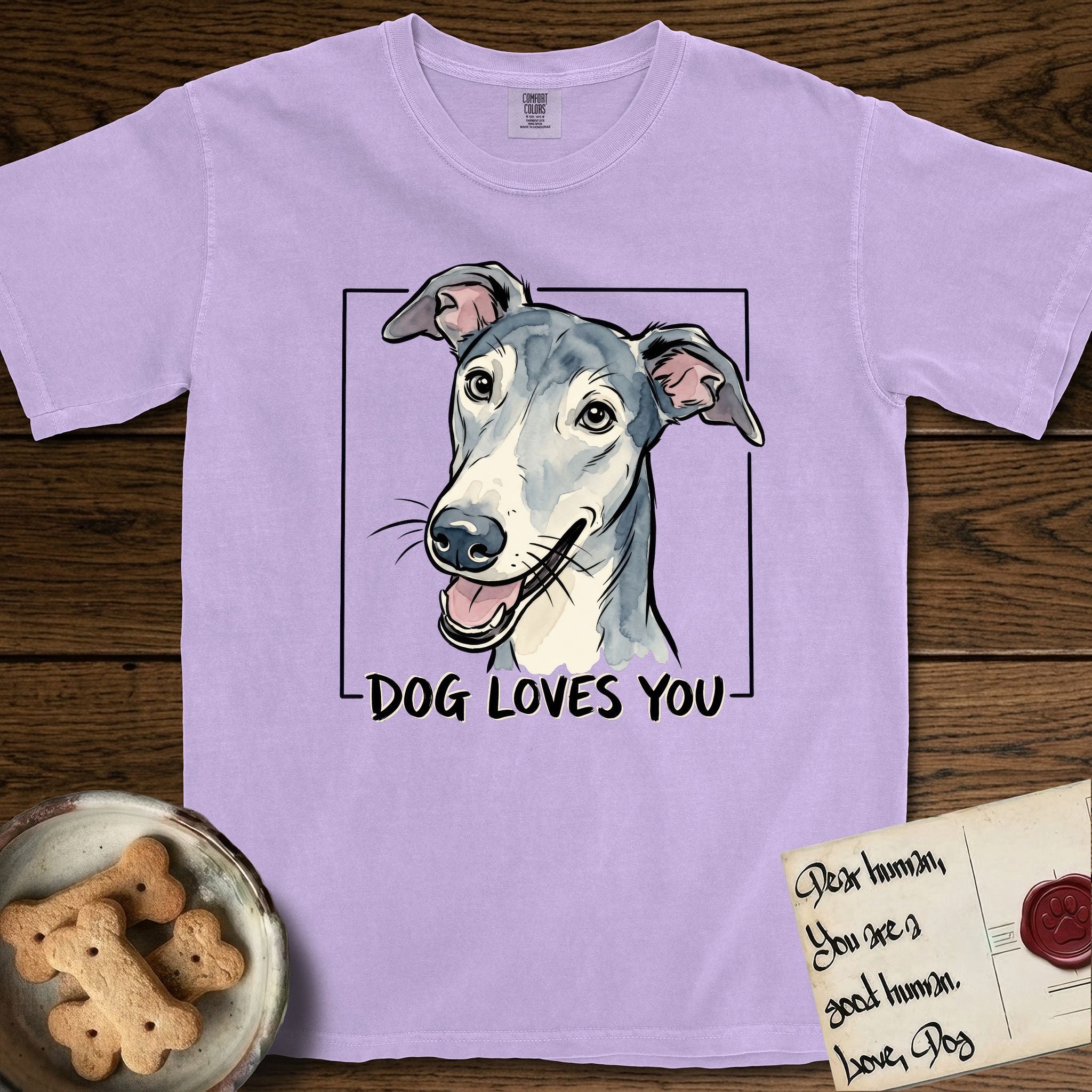 Dog Loves You Greyhound T-Shirt flat lay in Orchid, featuring Ink and watercolor portrait of a smiling slate blue Greyhound with a long narrow muzzle, cream blaze and folded rose ears, head tilted inside a black frame above hand-lettered text reading DOG LOVES YOU.