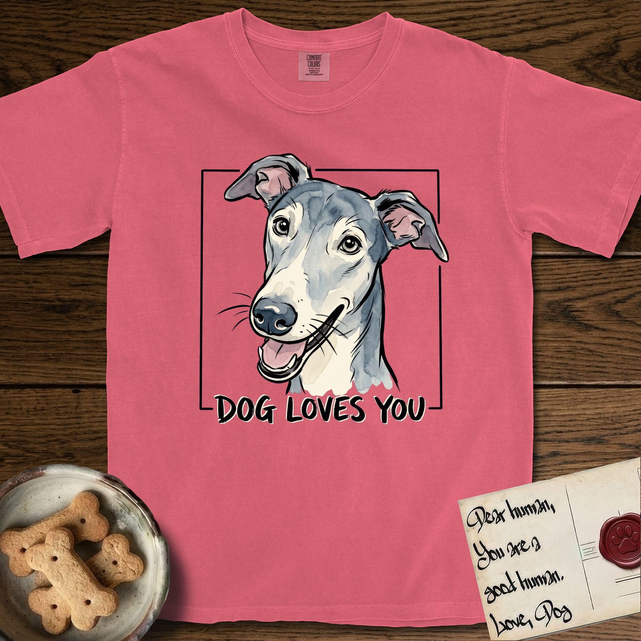 Dog Loves You Greyhound T-Shirt flat lay in Watermelon, featuring Ink and watercolor portrait of a smiling slate blue Greyhound with a long narrow muzzle, cream blaze and folded rose ears, head tilted inside a black frame above hand-lettered text reading DOG LOVES YOU.