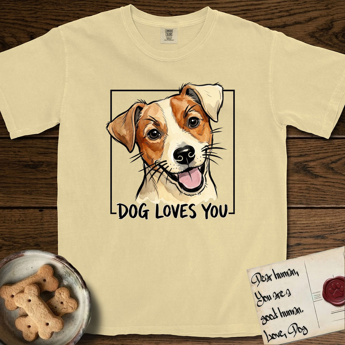 Dog Loves You Jack Russell T-Shirt flat lay in Butter, featuring Ink and watercolor portrait of a smiling cream and rust Jack Russell with folded button ears and keen dark eyes, head tilted inside a black frame above hand-lettered text reading DOG LOVES YOU.