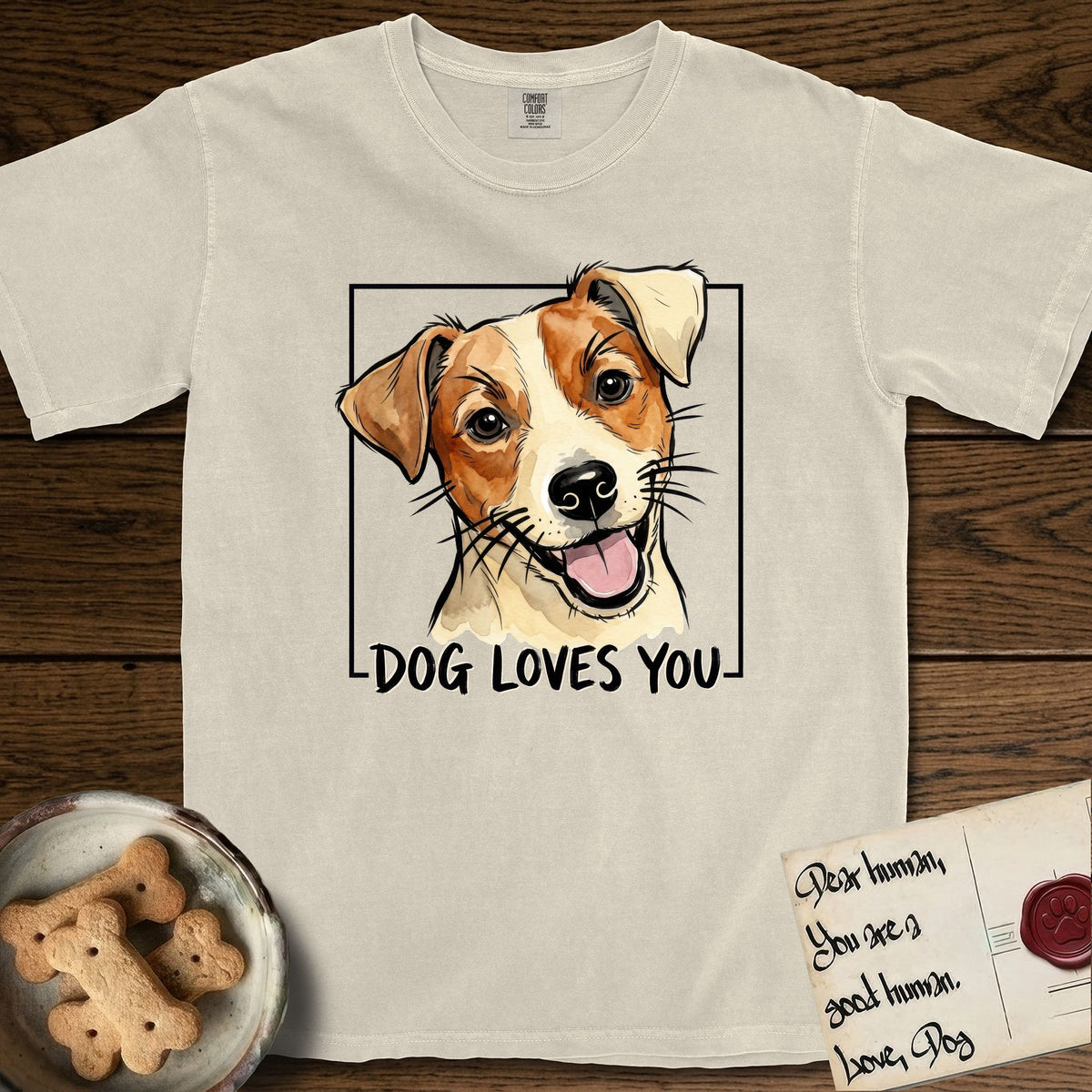 Dog Loves You Jack Russell T-Shirt flat lay in Ivory, featuring Ink and watercolor portrait of a smiling cream and rust Jack Russell with folded button ears and keen dark eyes, head tilted inside a black frame above hand-lettered text reading DOG LOVES YOU.