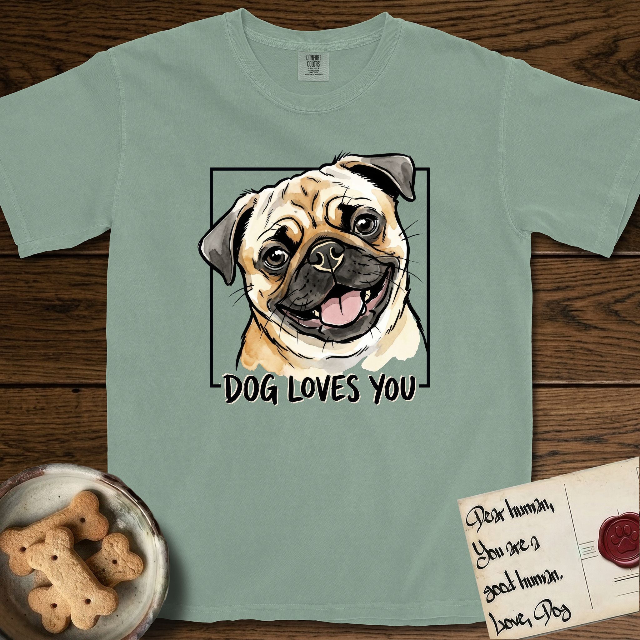 Dog Loves You Pug T-Shirt flat lay in Bay, featuring Ink and watercolor portrait of a smiling fawn Pug with a black mask, deep brow wrinkles and big round eyes, head tilted inside a black frame above hand-lettered text reading DOG LOVES YOU.