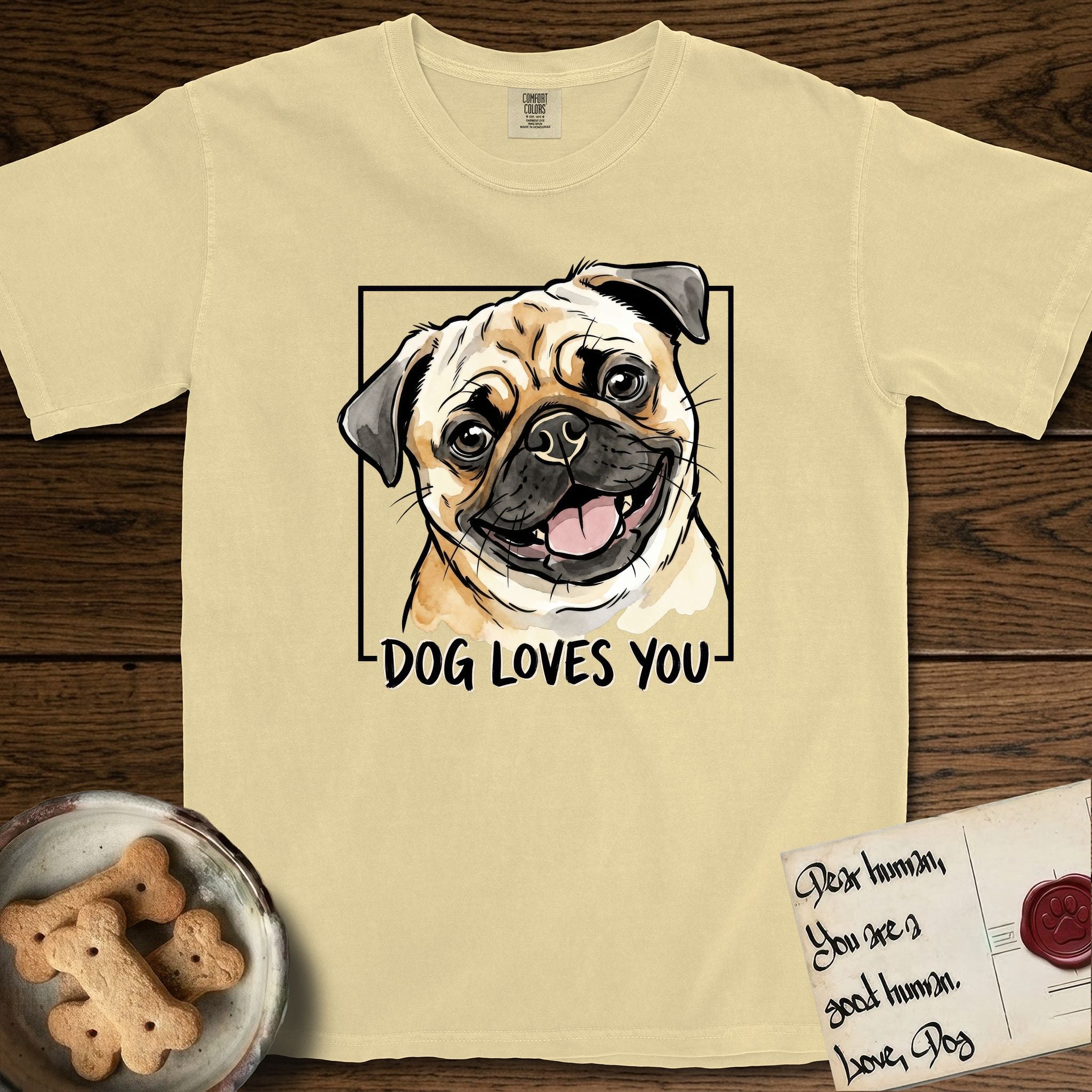 Dog Loves You Pug T-Shirt flat lay in Butter, featuring Ink and watercolor portrait of a smiling fawn Pug with a black mask, deep brow wrinkles and big round eyes, head tilted inside a black frame above hand-lettered text reading DOG LOVES YOU.