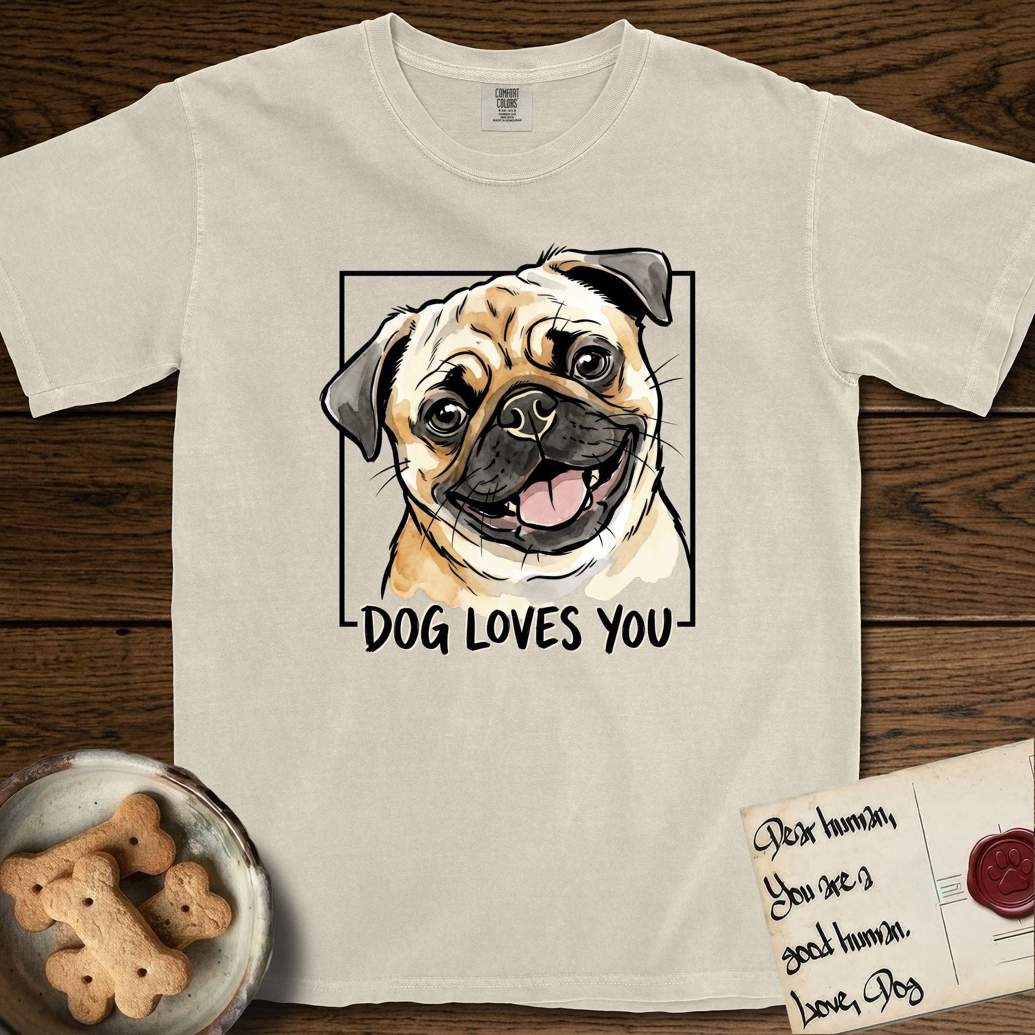 Dog Loves You Pug T-Shirt flat lay in Ivory, featuring Ink and watercolor portrait of a smiling fawn Pug with a black mask, deep brow wrinkles and big round eyes, head tilted inside a black frame above hand-lettered text reading DOG LOVES YOU.