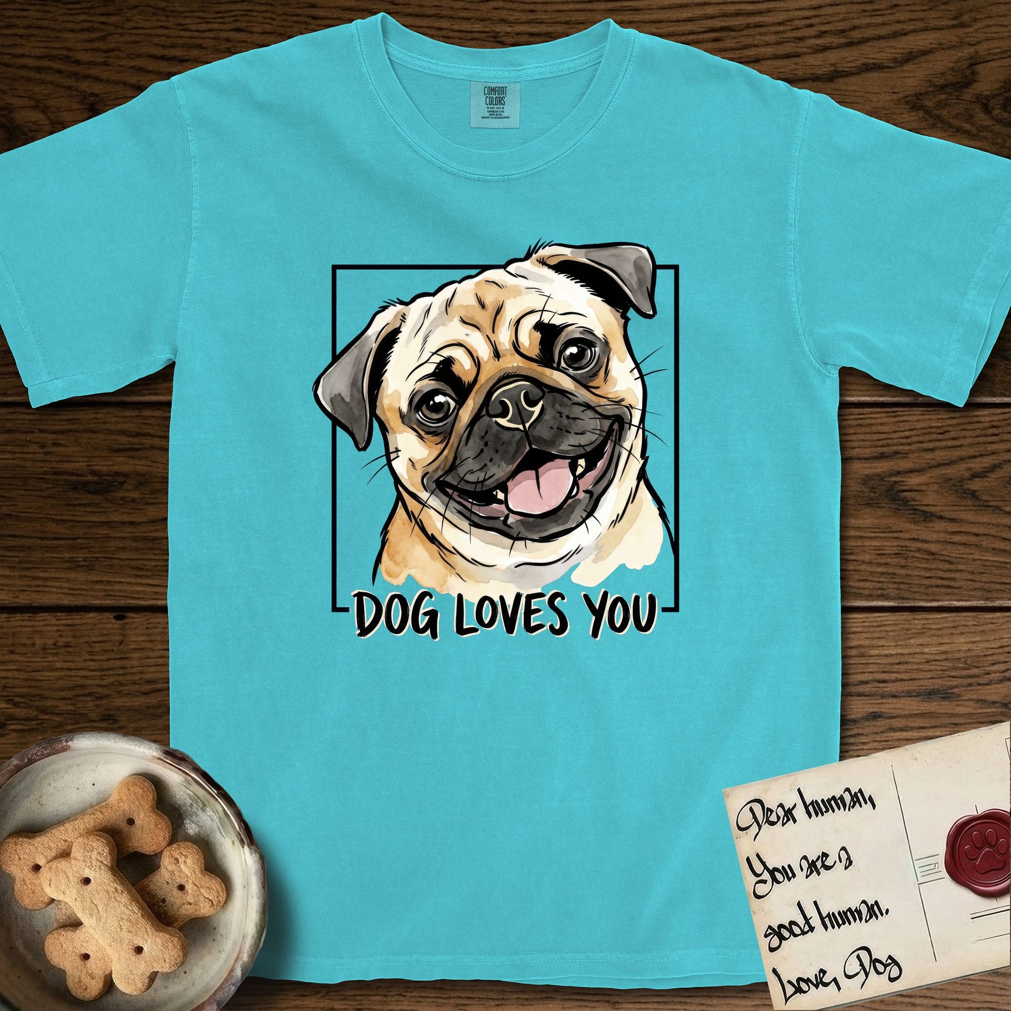 Dog Loves You Pug T-Shirt flat lay in Lagoon Blue, featuring Ink and watercolor portrait of a smiling fawn Pug with a black mask, deep brow wrinkles and big round eyes, head tilted inside a black frame above hand-lettered text reading DOG LOVES YOU.