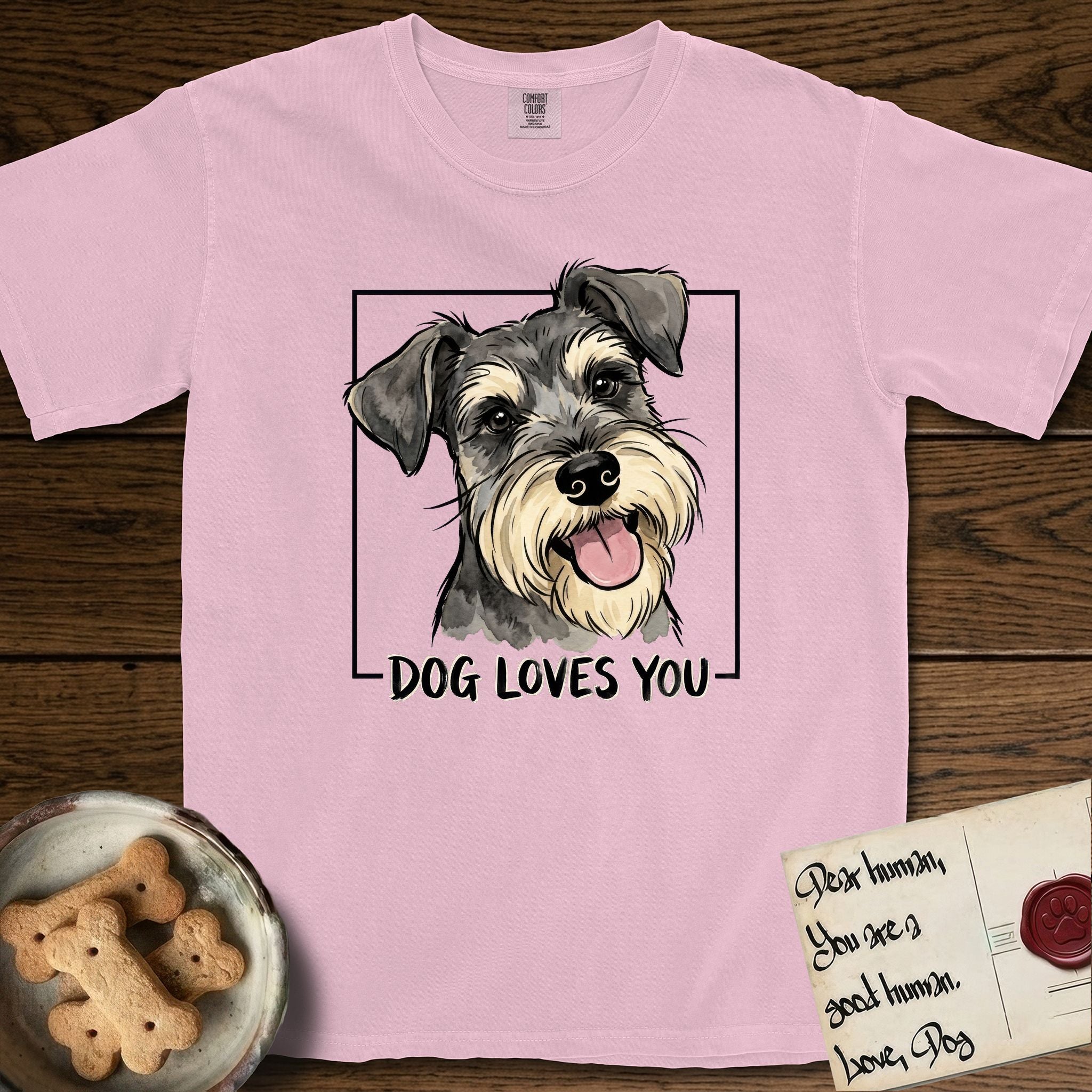 Dog Loves You Schnauzer T-Shirt flat lay in Blossom, featuring Ink and watercolor portrait of a smiling salt and pepper Schnauzer with a long beard and bushy eyebrows, head tilted inside a black frame above hand-lettered text reading DOG LOVES YOU.