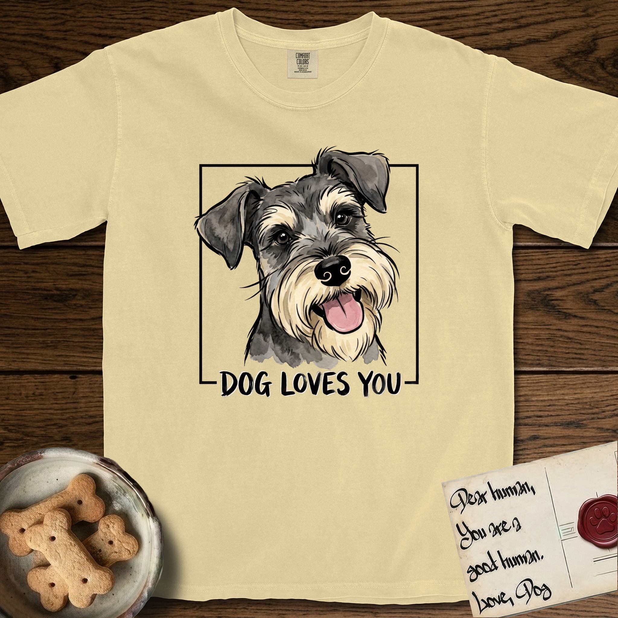 Dog Loves You Schnauzer T-Shirt flat lay in Butter, featuring Ink and watercolor portrait of a smiling salt and pepper Schnauzer with a long beard and bushy eyebrows, head tilted inside a black frame above hand-lettered text reading DOG LOVES YOU.