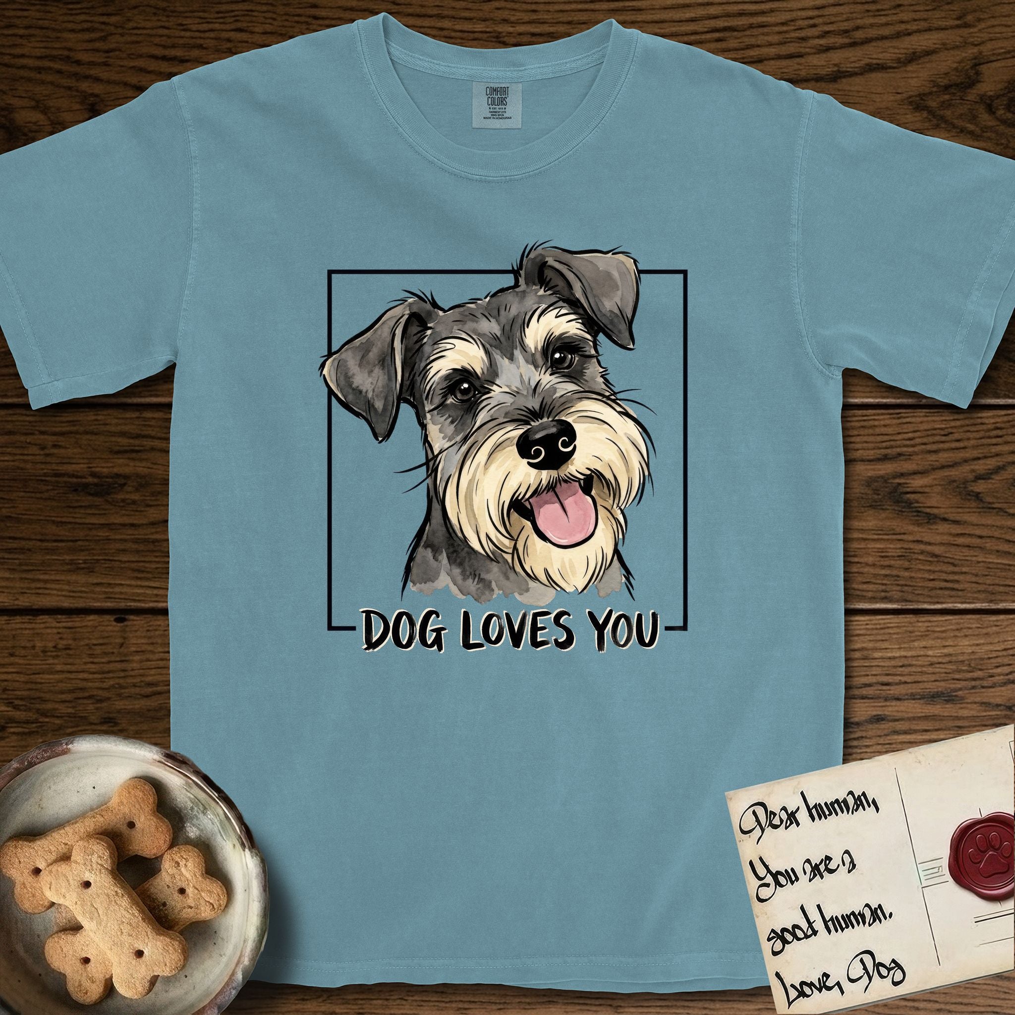 Dog Loves You Schnauzer T-Shirt flat lay in Ice Blue, featuring Ink and watercolor portrait of a smiling salt and pepper Schnauzer with a long beard and bushy eyebrows, head tilted inside a black frame above hand-lettered text reading DOG LOVES YOU.