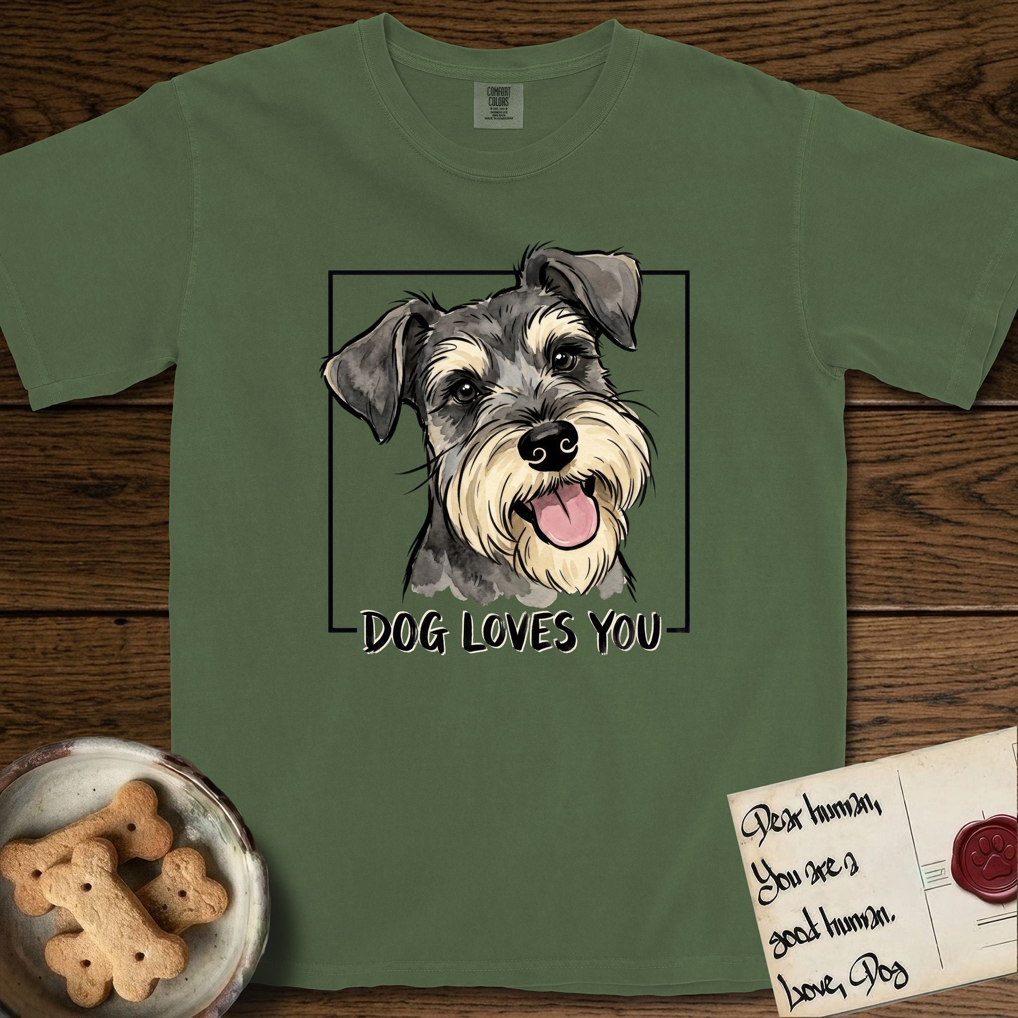 Dog Loves You Schnauzer T-Shirt flat lay in Moss, featuring Ink and watercolor portrait of a smiling salt and pepper Schnauzer with a long beard and bushy eyebrows, head tilted inside a black frame above hand-lettered text reading DOG LOVES YOU.