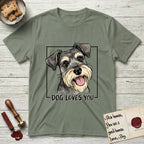 Dog Loves You Schnauzer T-Shirt flat lay in Sage, featuring Ink and watercolor portrait of a smiling salt and pepper Schnauzer with a long beard and bushy eyebrows, head tilted inside a black frame above hand-lettered text reading DOG LOVES YOU.
