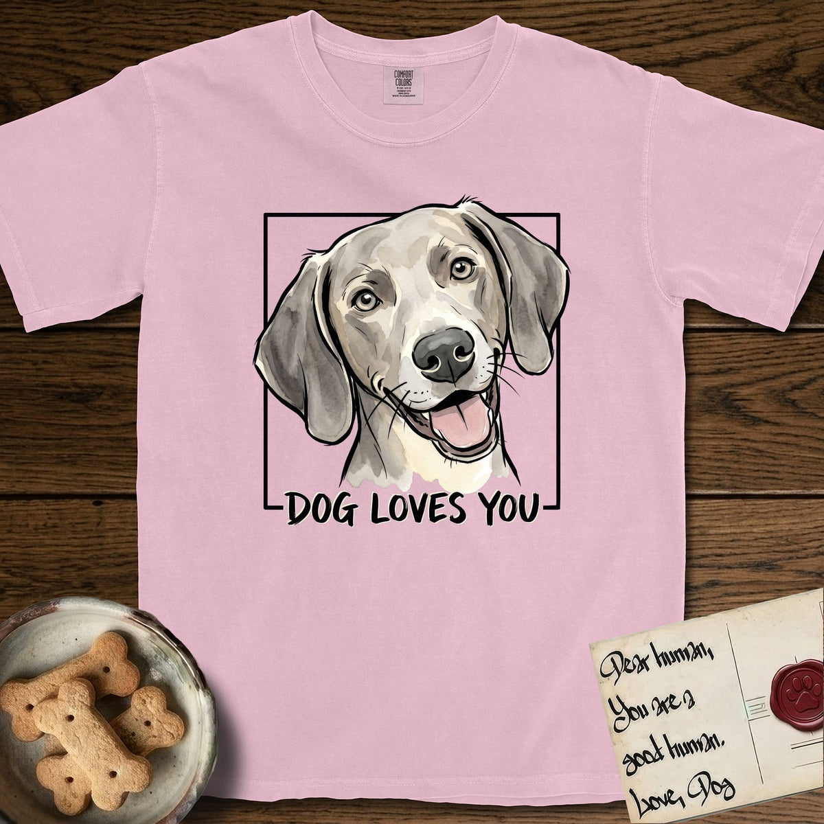 Dog Loves You Weimaraner T-Shirt flat lay in Blossom, featuring Ink and watercolor portrait of a smiling silver grey Weimaraner with long velvet hanging ears and pale amber eyes, head tilted inside a black frame above hand-lettered text reading DOG LOVES YOU.