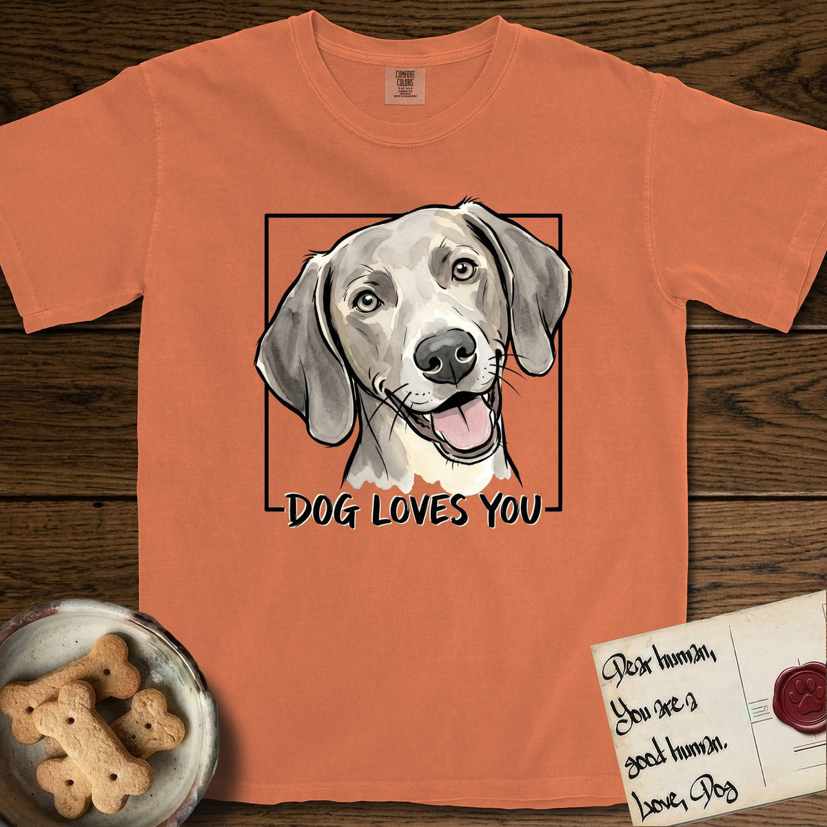 Dog Loves You Weimaraner T-Shirt flat lay in Melon, featuring Ink and watercolor portrait of a smiling silver grey Weimaraner with long velvet hanging ears and pale amber eyes, head tilted inside a black frame above hand-lettered text reading DOG LOVES YOU.