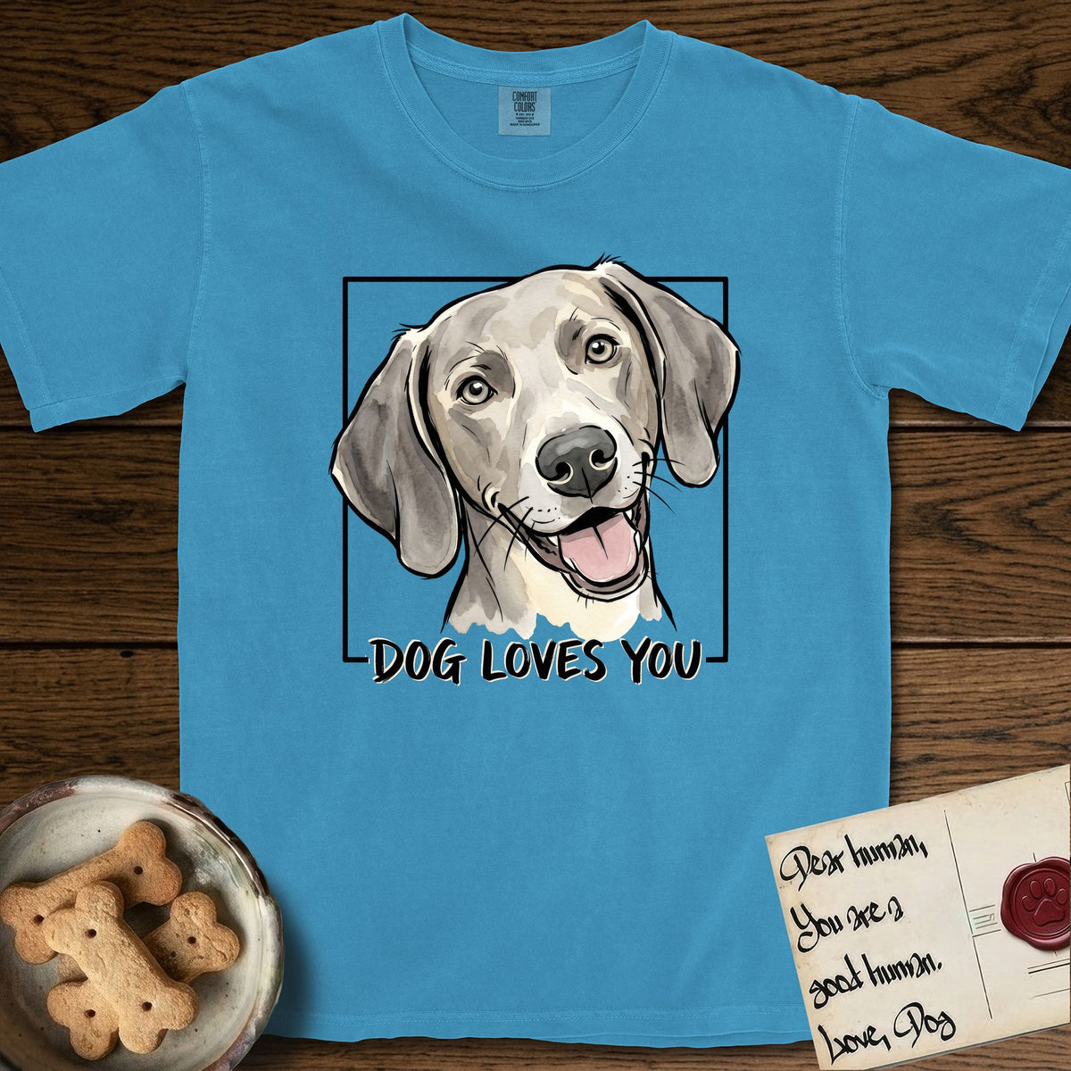 Dog Loves You Weimaraner T-Shirt flat lay in Royal Caribe, featuring Ink and watercolor portrait of a smiling silver grey Weimaraner with long velvet hanging ears and pale amber eyes, head tilted inside a black frame above hand-lettered text reading DOG LOVES YOU.