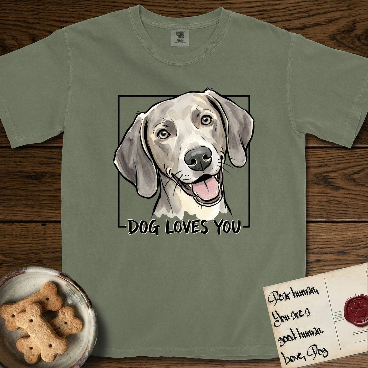 Dog Loves You Weimaraner T-Shirt flat lay in Sage, featuring Ink and watercolor portrait of a smiling silver grey Weimaraner with long velvet hanging ears and pale amber eyes, head tilted inside a black frame above hand-lettered text reading DOG LOVES YOU.