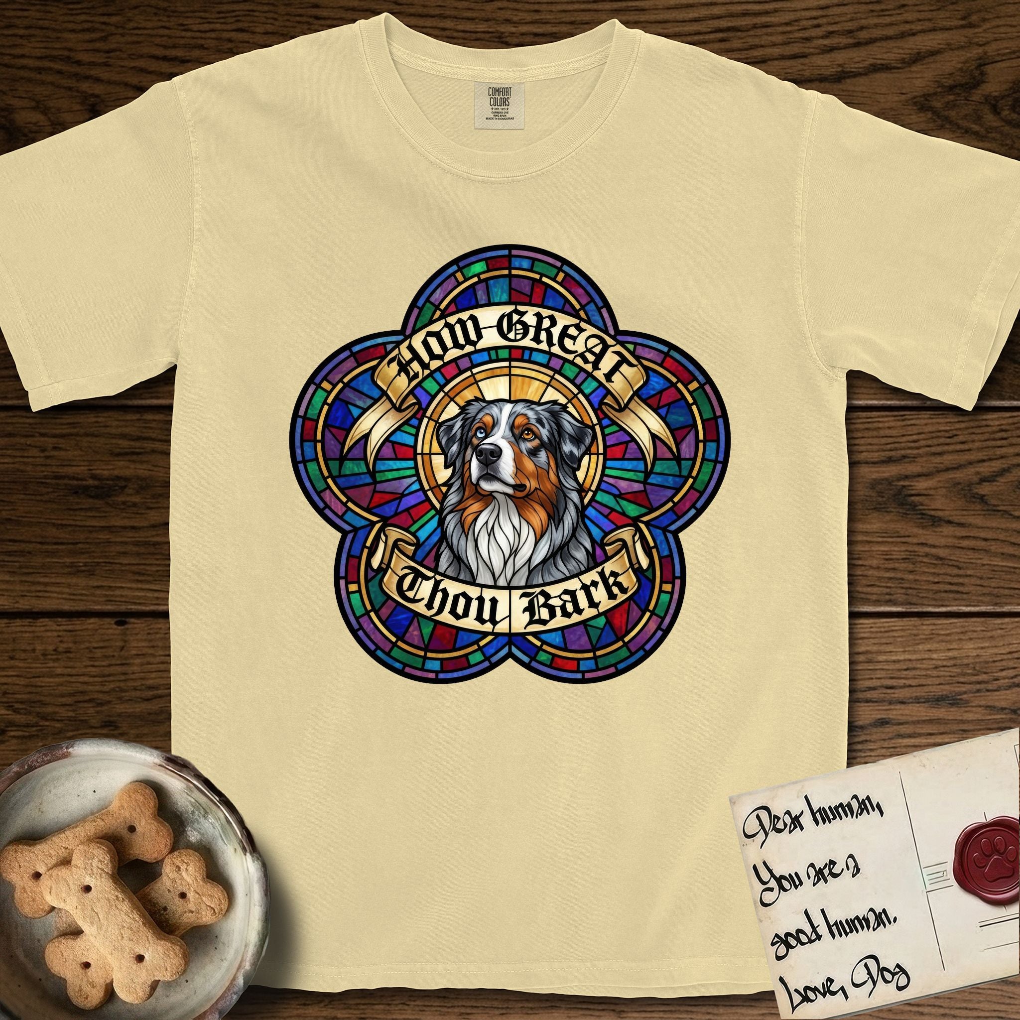 How Great Thou Bark Australian Shepherd T-Shirt flat lay in Butter, featuring a full-color stained-glass saint window of a blue merle Australian Shepherd with one blue eye and one amber eye, haloed in golden light.