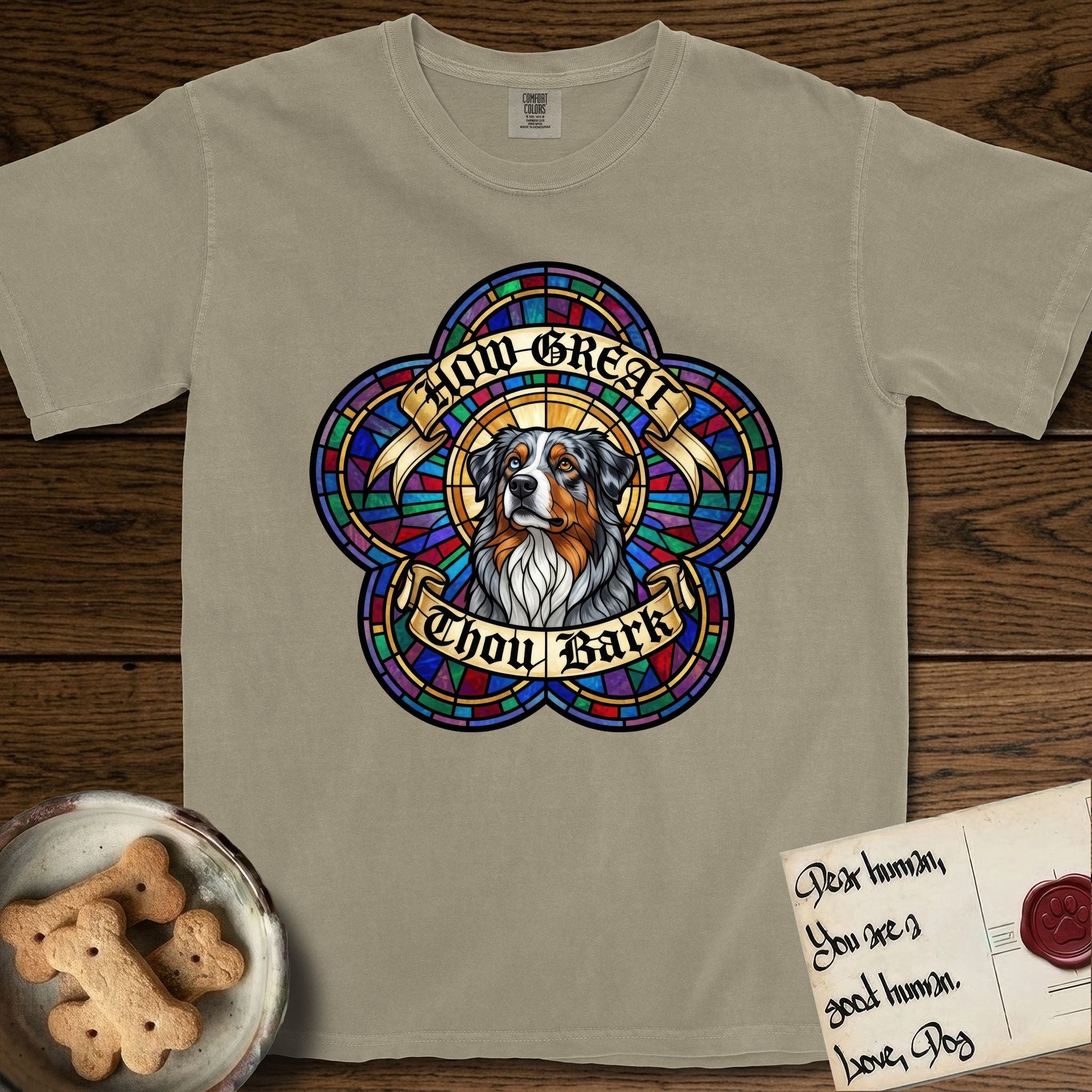 How Great Thou Bark Australian Shepherd T-Shirt flat lay in Khaki, featuring a full-color stained-glass saint window of a blue merle Australian Shepherd with one blue eye and one amber eye, haloed in golden light.