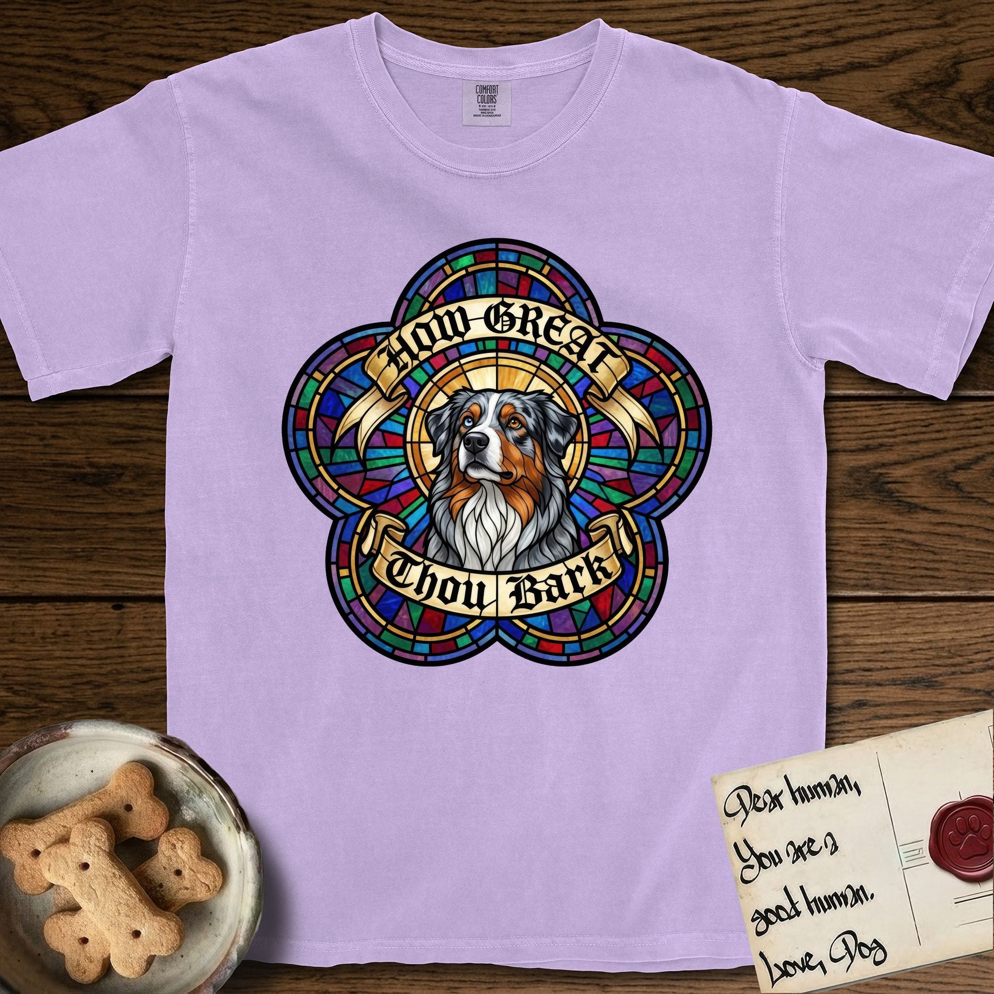 How Great Thou Bark Australian Shepherd T-Shirt flat lay in Orchid, featuring a full-color stained-glass saint window of a blue merle Australian Shepherd with one blue eye and one amber eye, haloed in golden light.