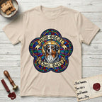How Great Thou Bark Australian Shepherd T-Shirt flat lay in Sand, featuring a full-color stained-glass saint window of a blue merle Australian Shepherd with one blue eye and one amber eye, haloed in golden light.