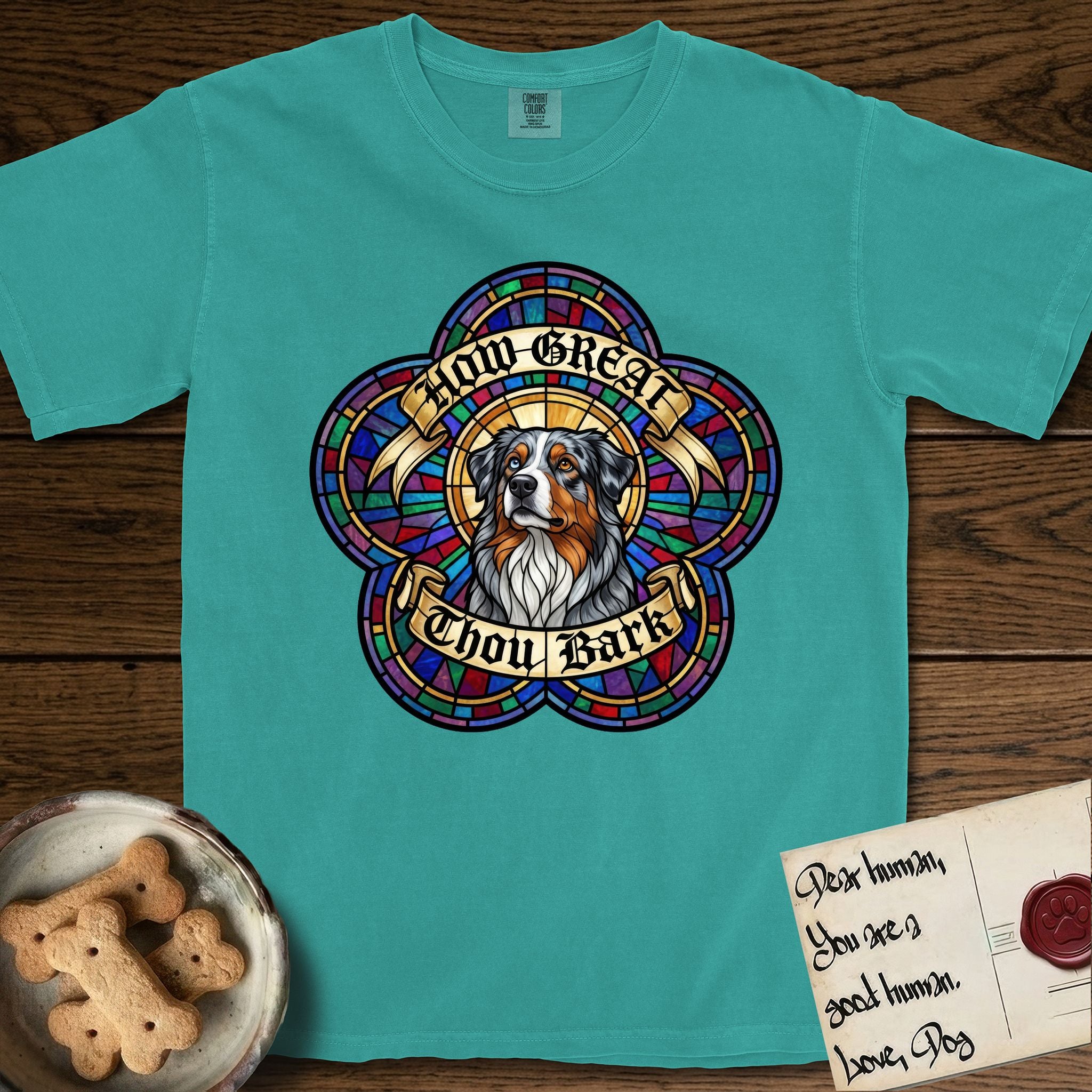 How Great Thou Bark Australian Shepherd T-Shirt flat lay in Seafoam, featuring a full-color stained-glass saint window of a blue merle Australian Shepherd with one blue eye and one amber eye, haloed in golden light.