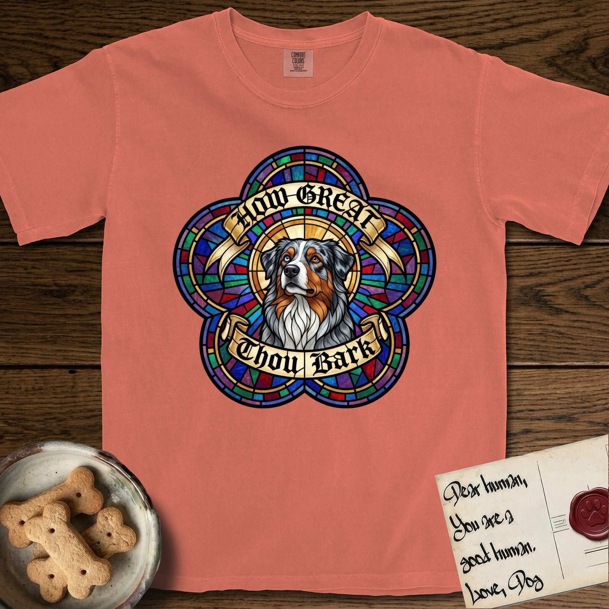 How Great Thou Bark Australian Shepherd T-Shirt flat lay in Terracotta, featuring a full-color stained-glass saint window of a blue merle Australian Shepherd with one blue eye and one amber eye, haloed in golden light.