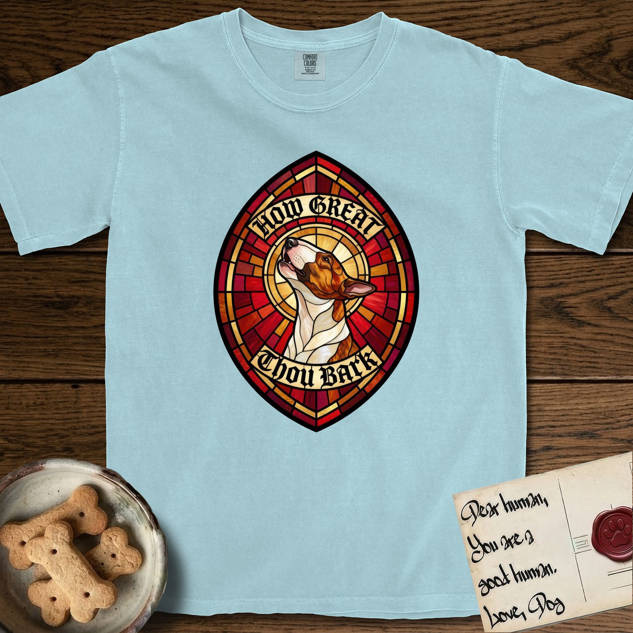 How Great Thou Bark Bull Terrier T-Shirt flat lay in Chambray, featuring a full-color stained-glass saint window of a brown and white Bull Terrier with head raised and mouth open mid-howl, haloed in golden light.